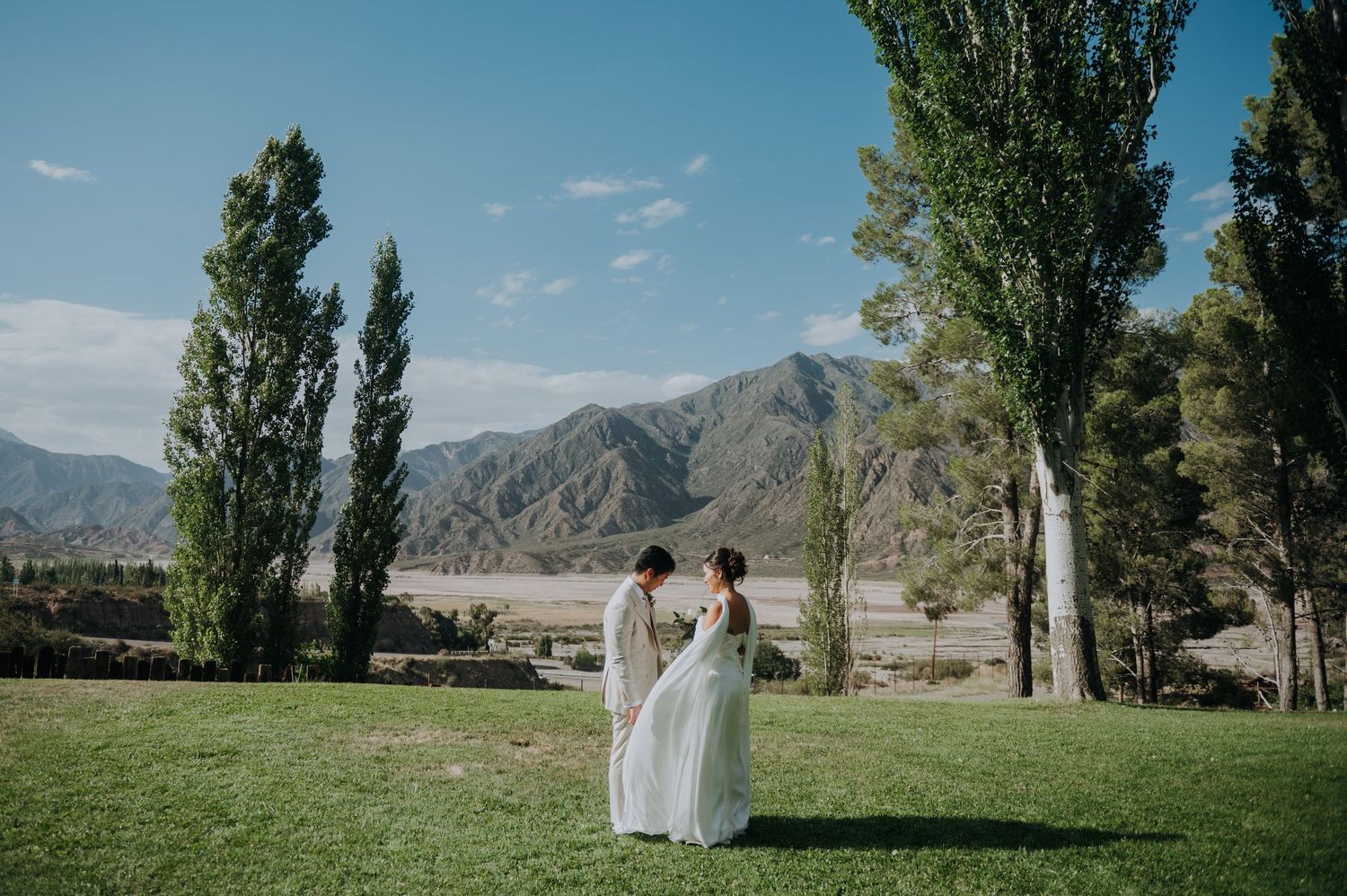 Bride and groom share a romantic moment on green lawn with tall trees and mountain backdrop under blue sky.