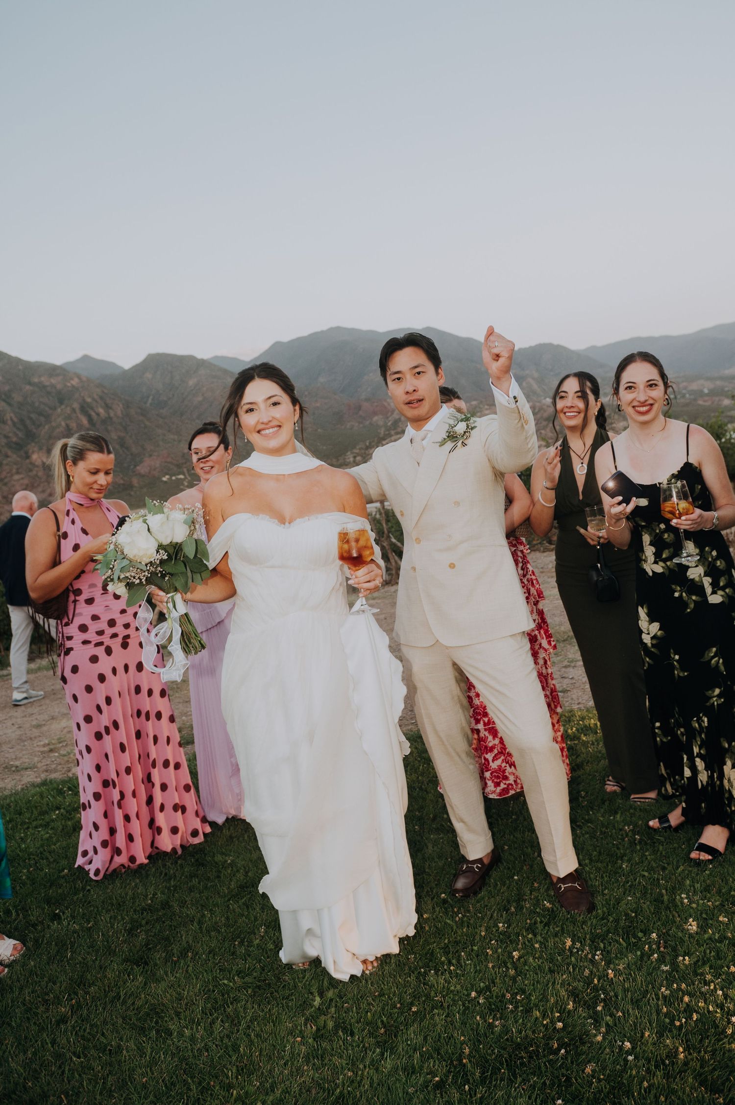 Bride and groom celebrate outdoors with wedding guests in colorful dresses against a scenic mountain backdrop.