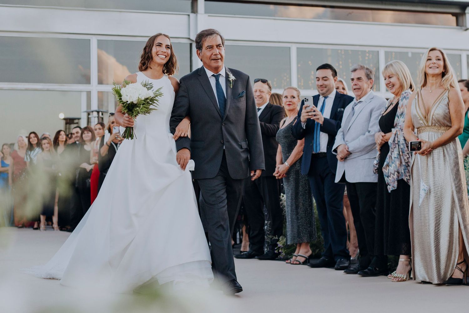 Bride in white gown walks down aisle with older man in suit, guests watching at elegant wedding venue.