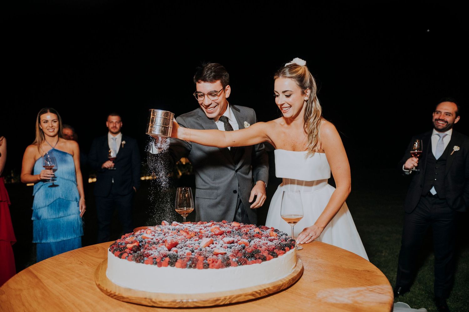 Bride and groom cutting a large berry-topped wedding cake outdoors at night with guests watching.