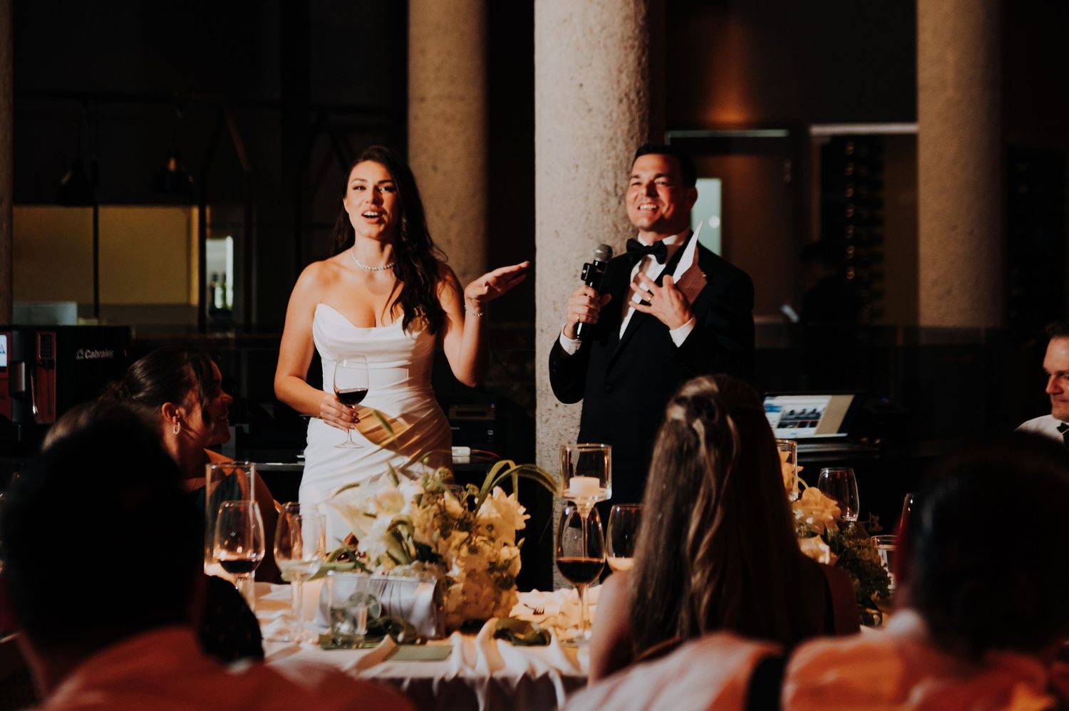 Bride and groom giving speech at elegant candlelit wedding reception with floral centerpieces and seated guests.
