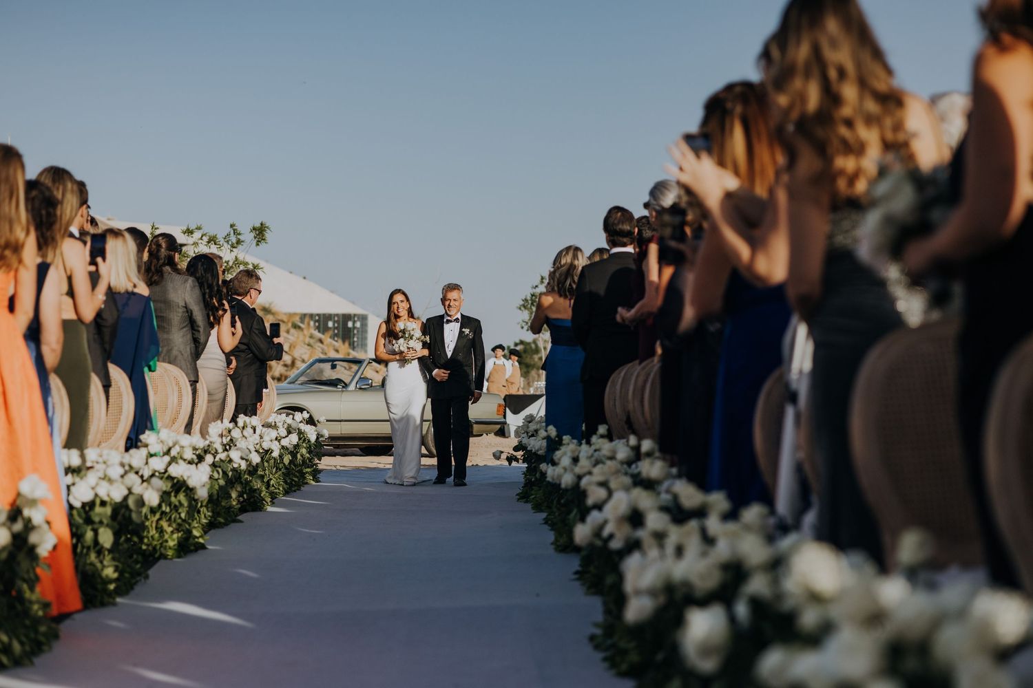 Bride and groom walking down outdoor aisle lined with white flowers and guests.