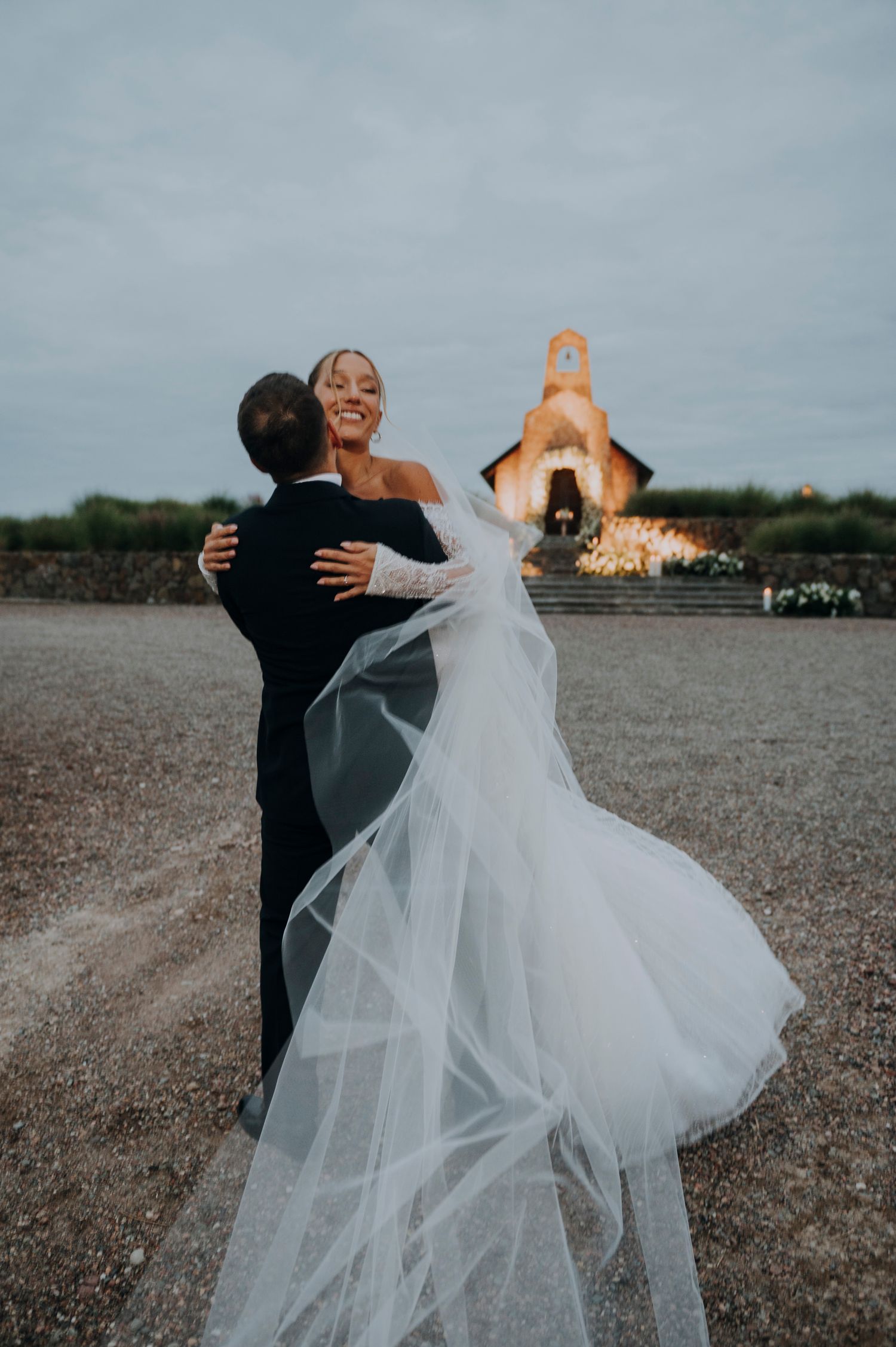 Groom lifts bride in flowing white veil outside rustic chapel at dusk.