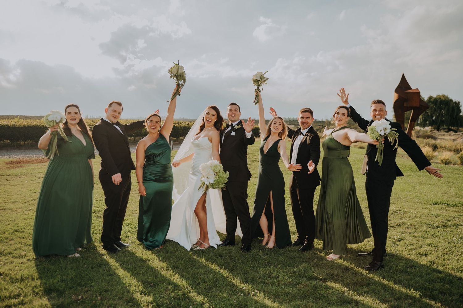 Wedding party celebrating outdoors in green and black attire with bride in white gown raising bouquet on lush lawn.