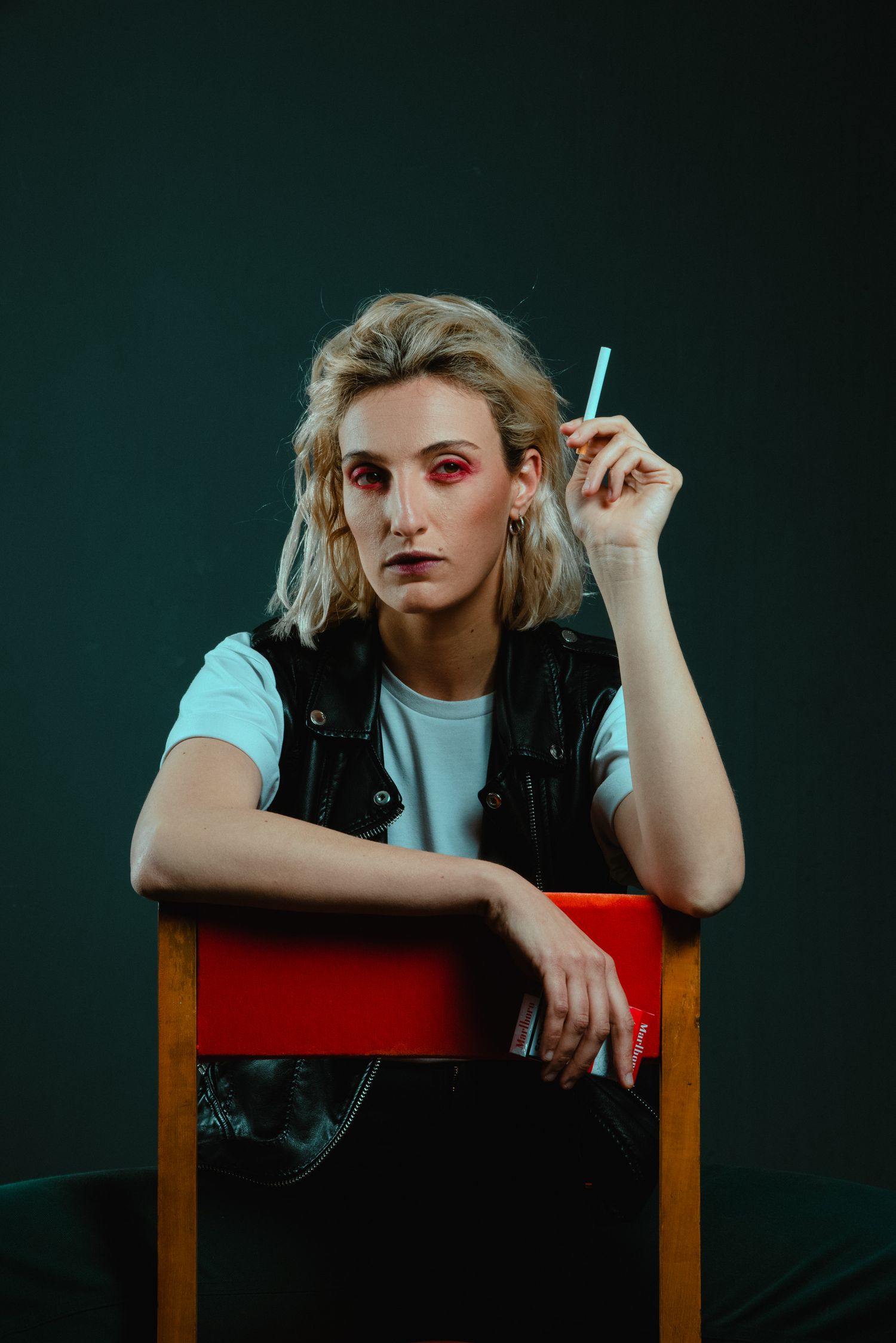 Blonde woman with bold red eye makeup sitting on a chair holding a pen against dark teal background.