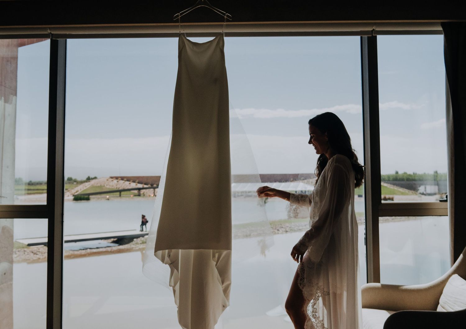 Bride in sheer robe standing by large window with wedding dress hanging, overlooking scenic waterfront view.