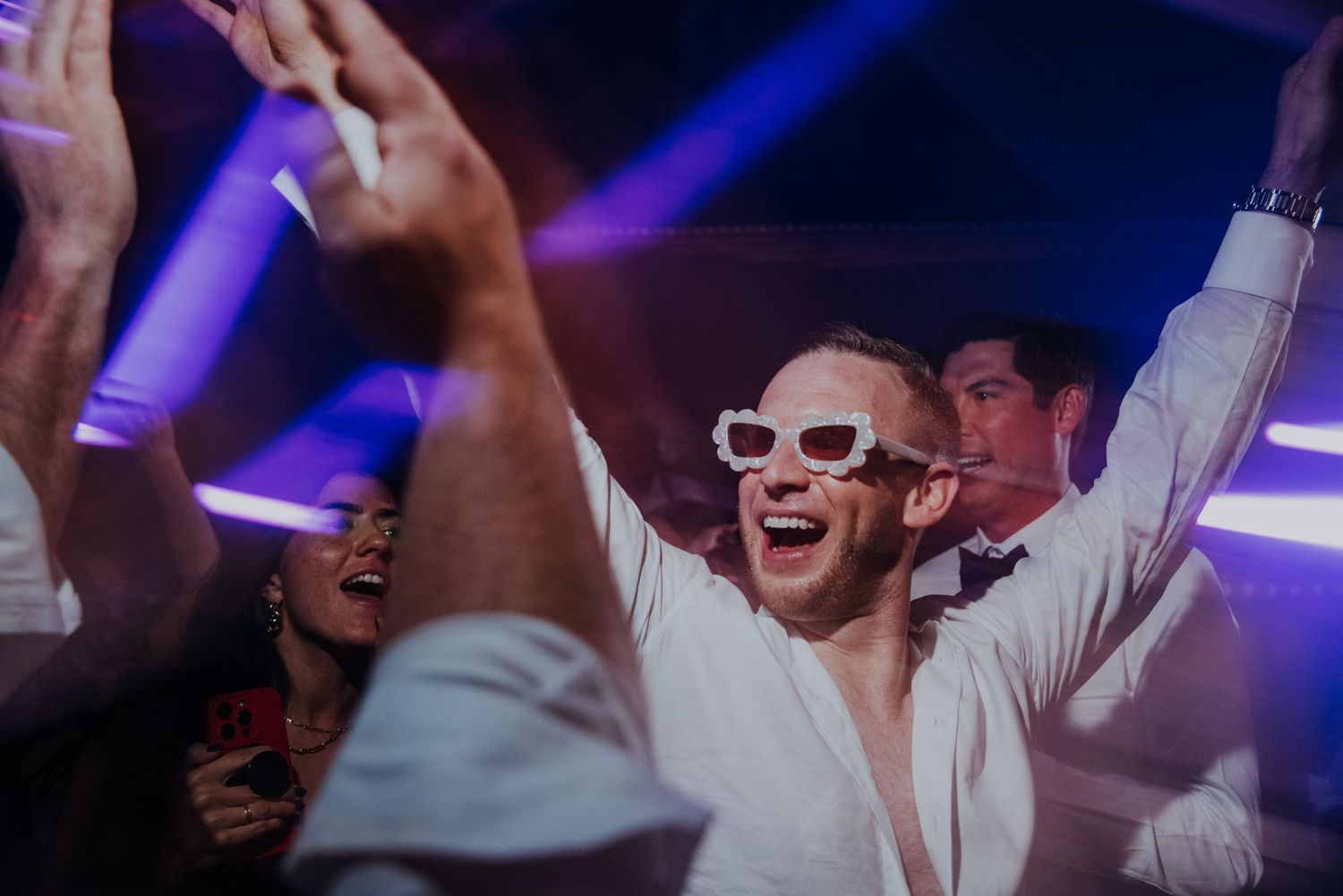 Groom celebrates on the dance floor wearing white sunglasses at a wedding reception.