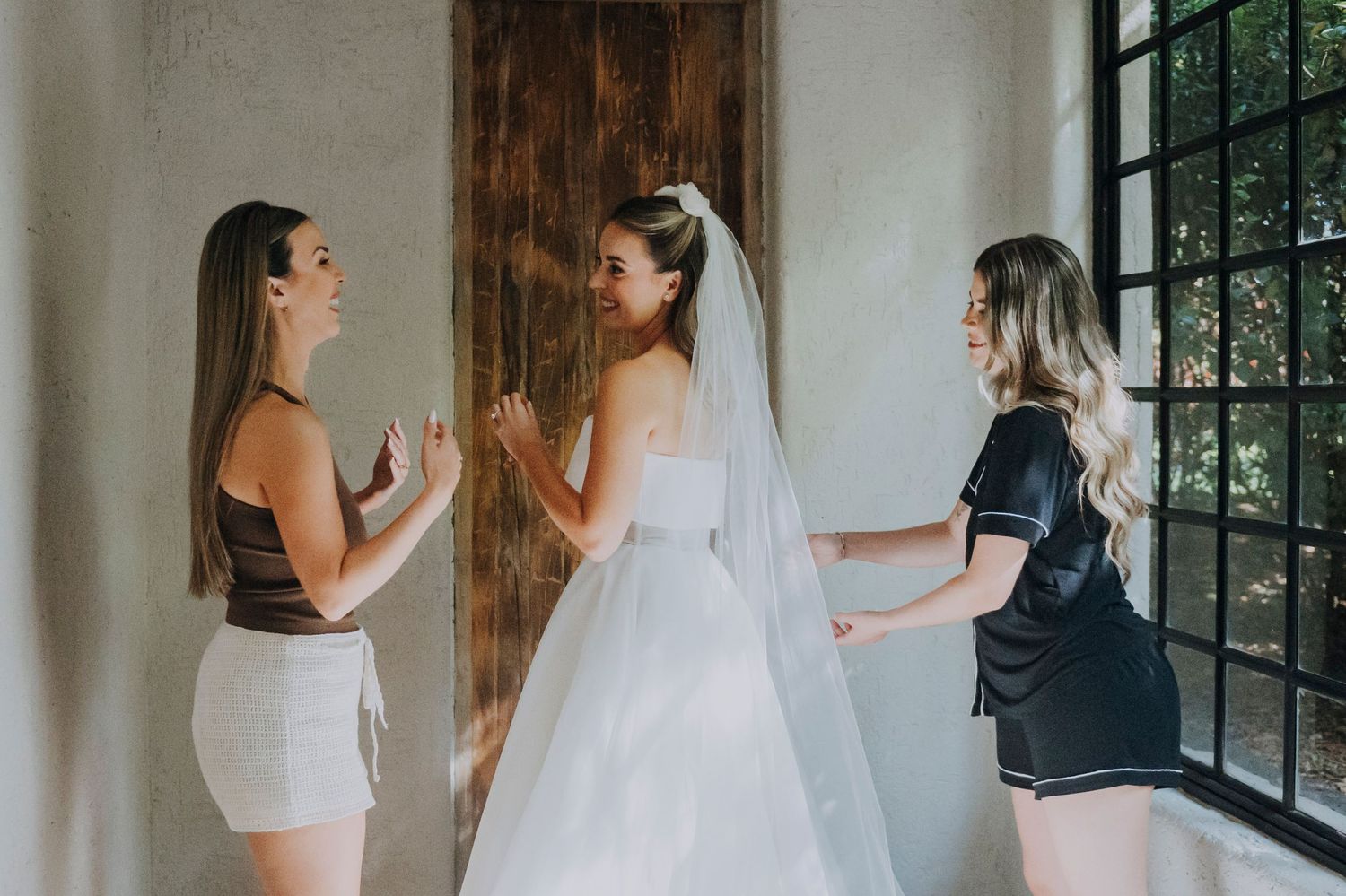 Bride in white gown and veil with two bridesmaids in casual attire helping her prepare indoors.