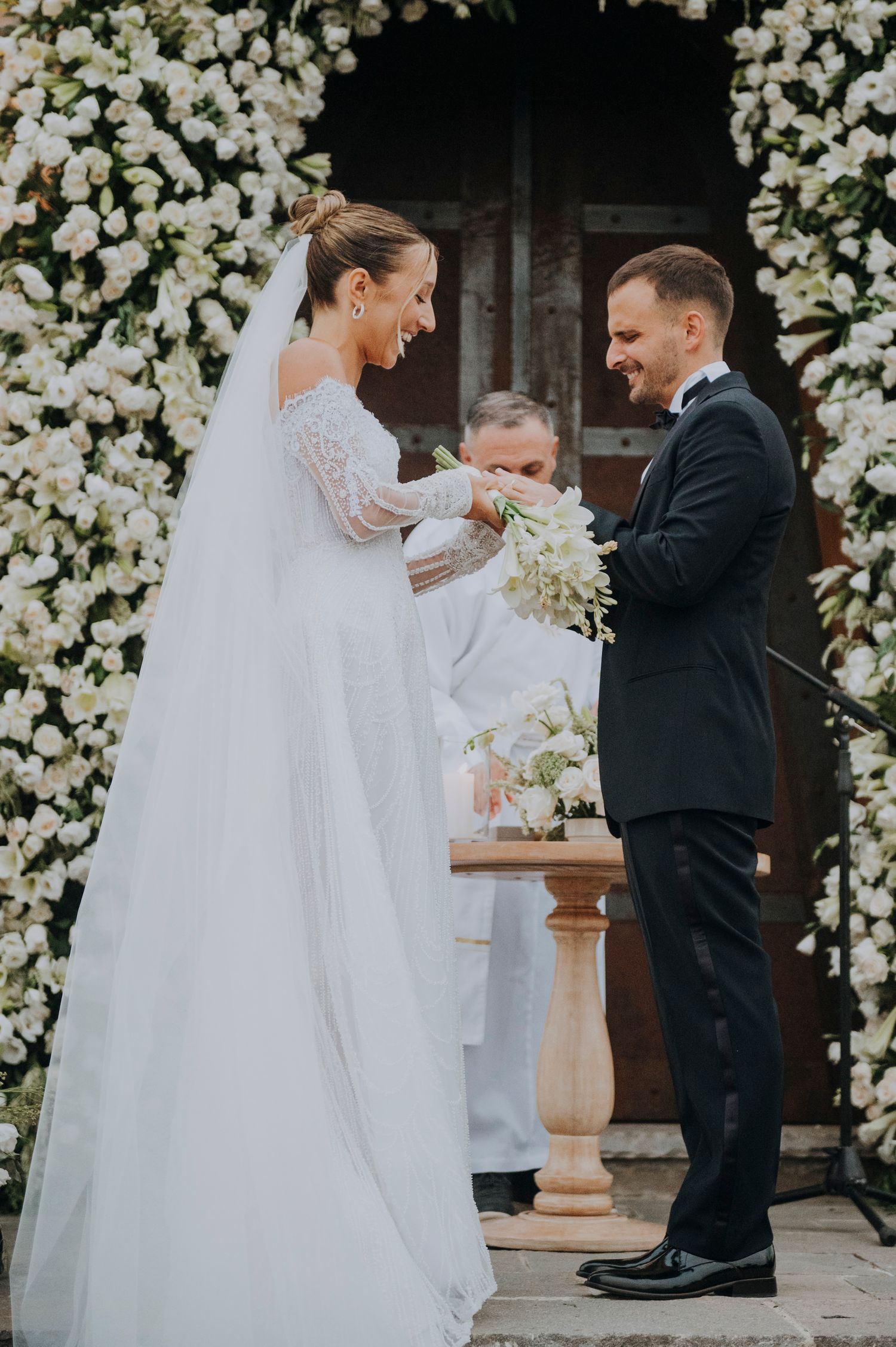 Bride and groom exchanging rings at outdoor wedding ceremony with white floral arch backdrop.