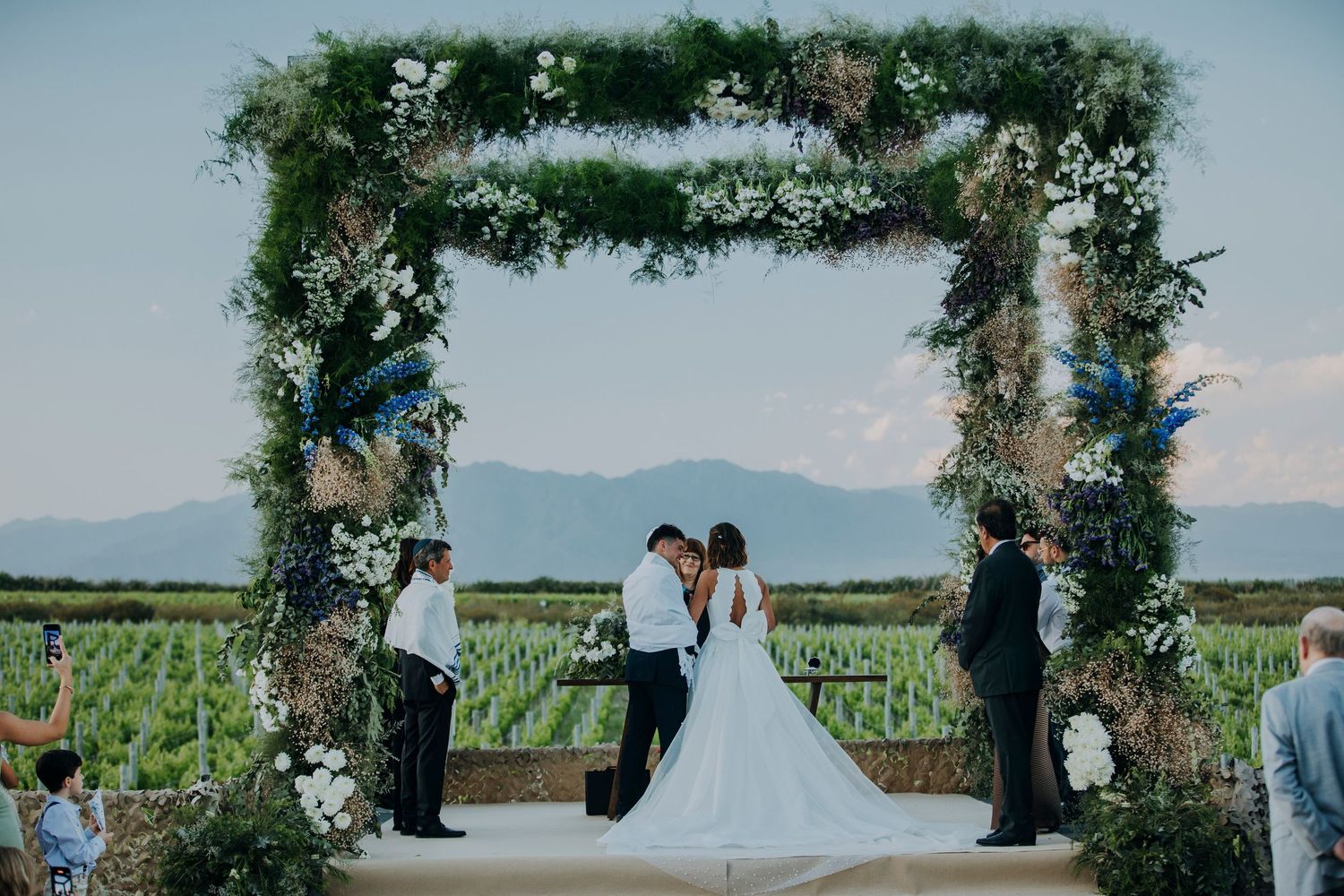 Bride and groom exchange vows under a floral arch at a vineyard wedding with mountain views.