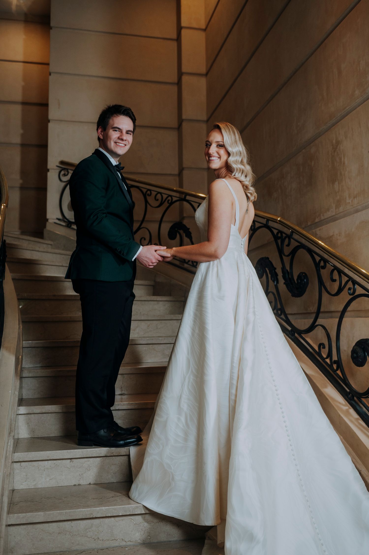 Bride in white ballgown and groom in green tuxedo holding hands on staircase.