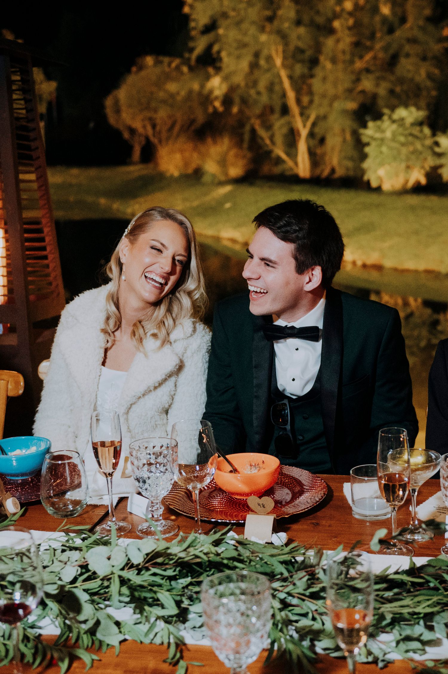 Laughing bride and groom sit together at candlelit outdoor wedding reception dinner table at night.
