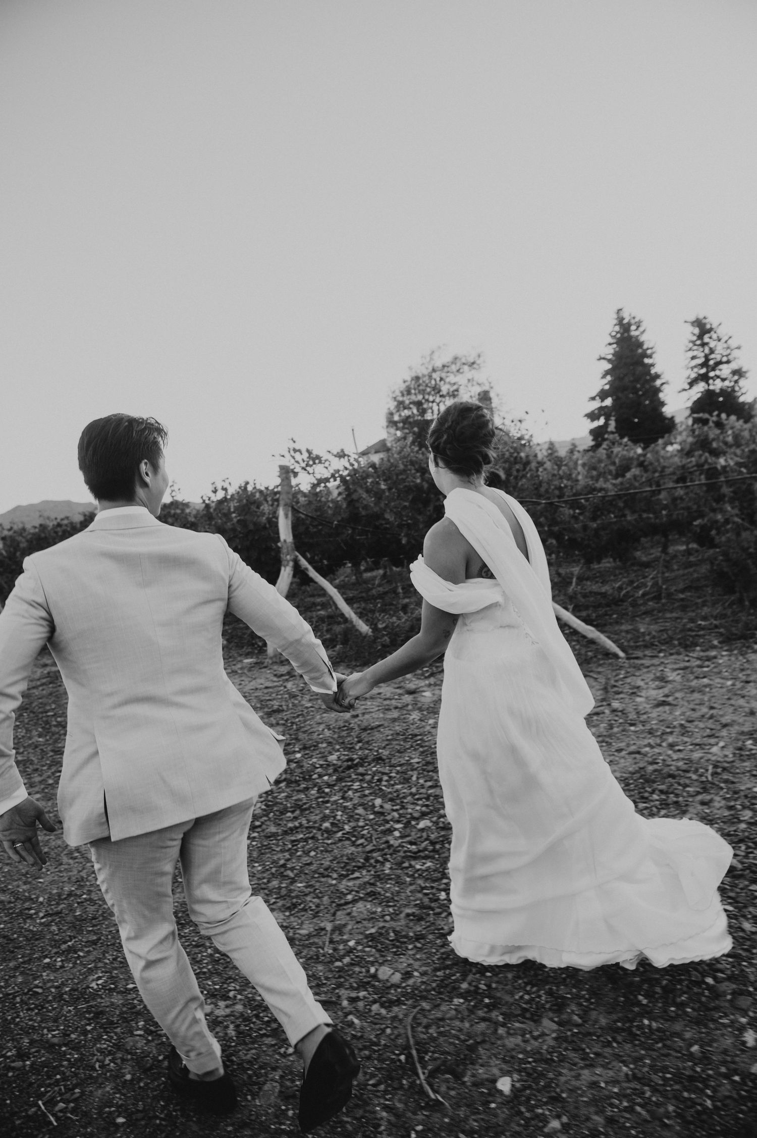 Black and white photo of bride and groom running hand-in-hand through vineyard after wedding ceremony.
