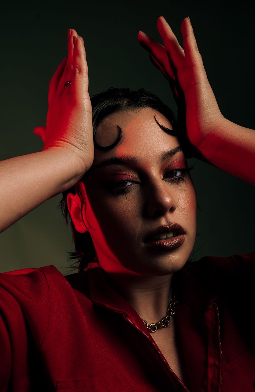GUGA VALDEZ Woman in red outfit and gloves with dramatic makeup poses with hands raised above head against dark background.