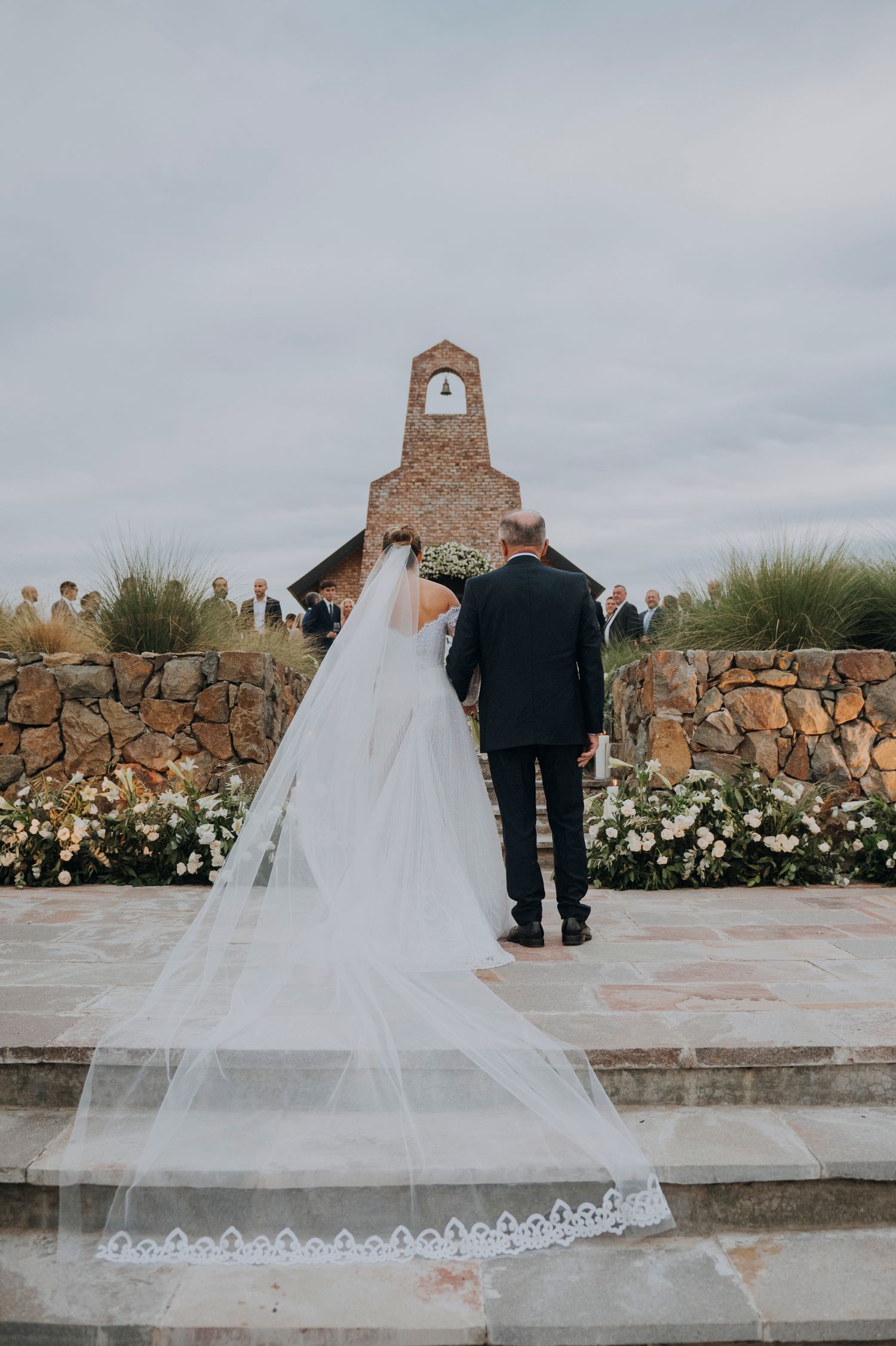 Bride in flowing white gown with long lace-trimmed veil walks toward rustic stone chapel altar with escort.