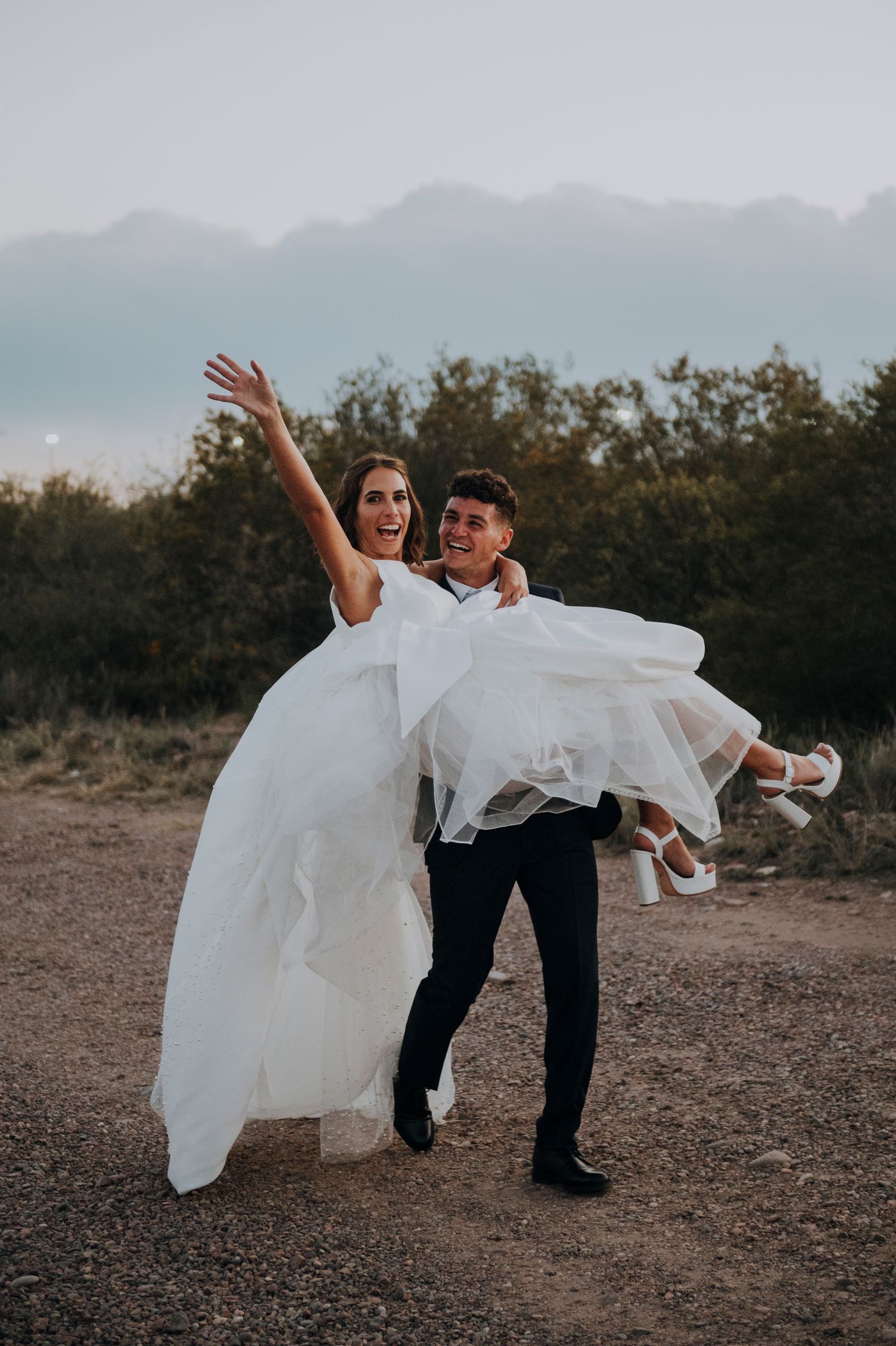 Joyful groom carries laughing bride in white wedding gown outdoors with mountains in background.