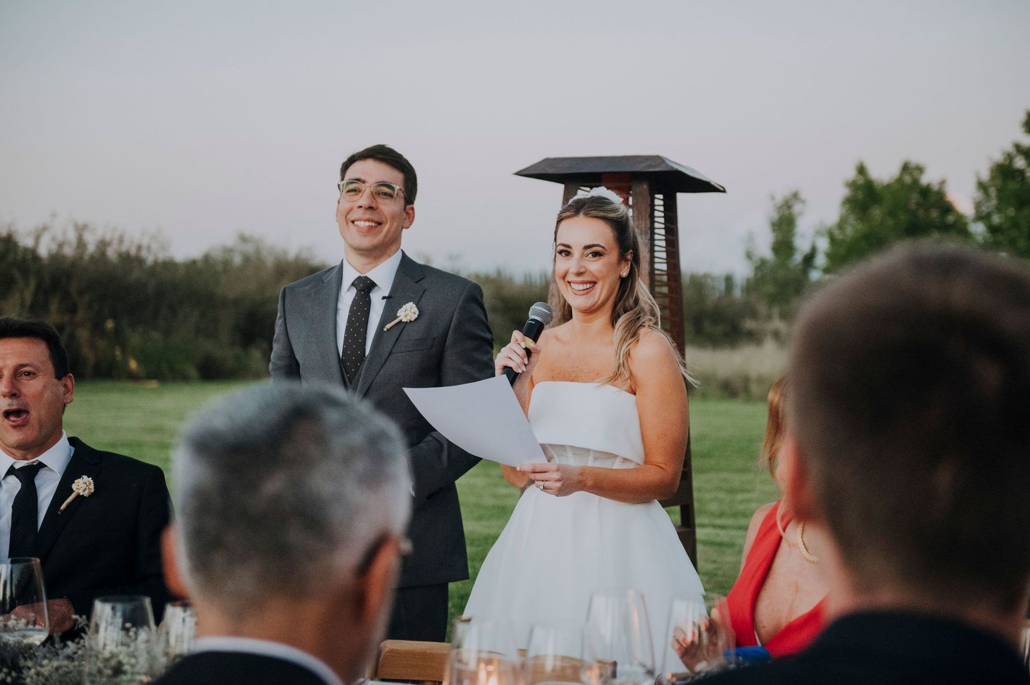 Smiling bride speaks into microphone at outdoor reception while groom laughs beside her.