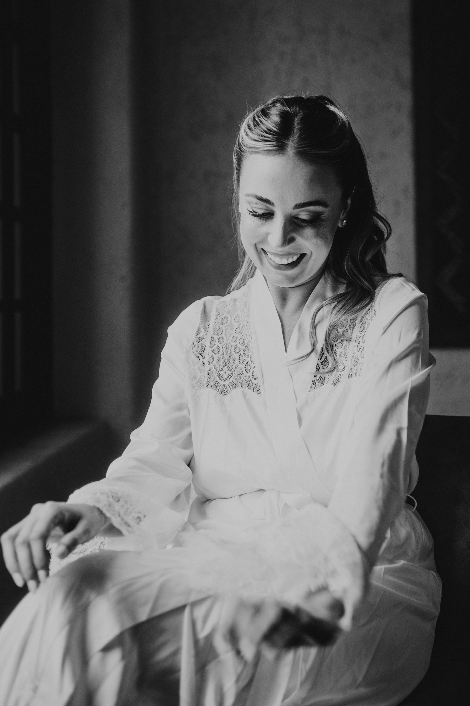 Smiling bride in a white lace robe sitting by a window in black and white wedding preparation photo.