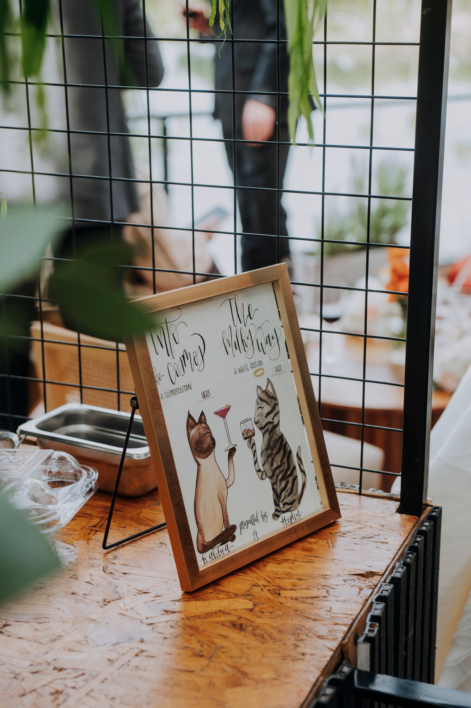 Framed cat-illustrated wedding cocktail menu sign displayed on wooden surface at wedding bar station.