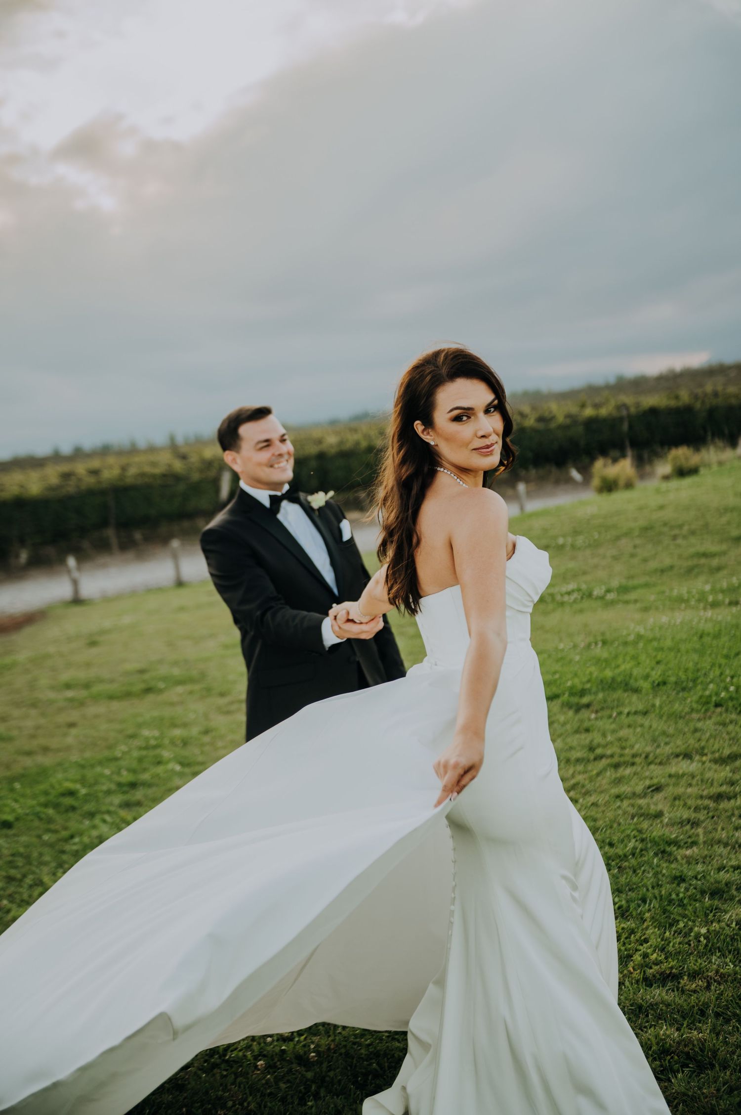 Bride in strapless gown twirling with groom in tuxedo on green lawn at dusk.