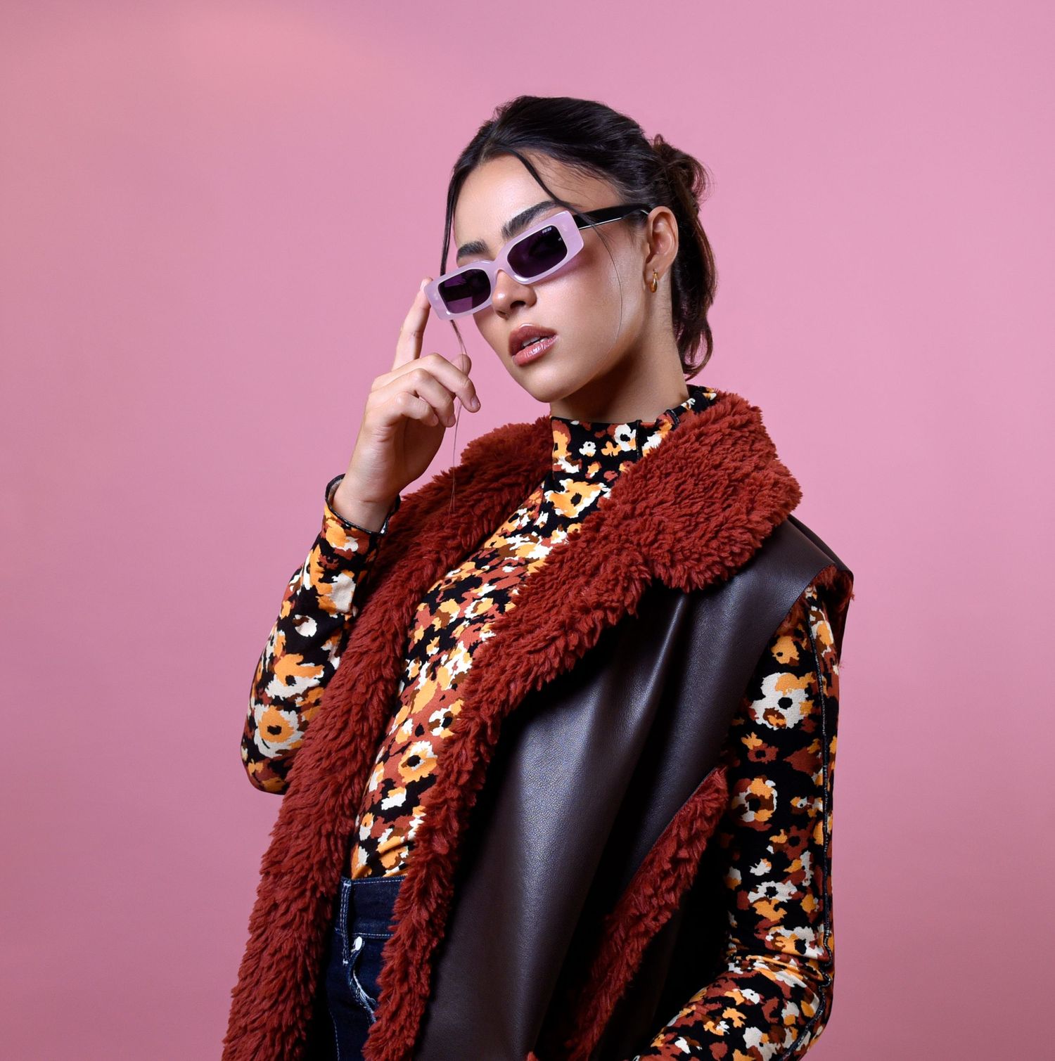 Stylish Asian woman in floral top, leather vest and rust sherpa coat adjusting sunglasses on pink background.