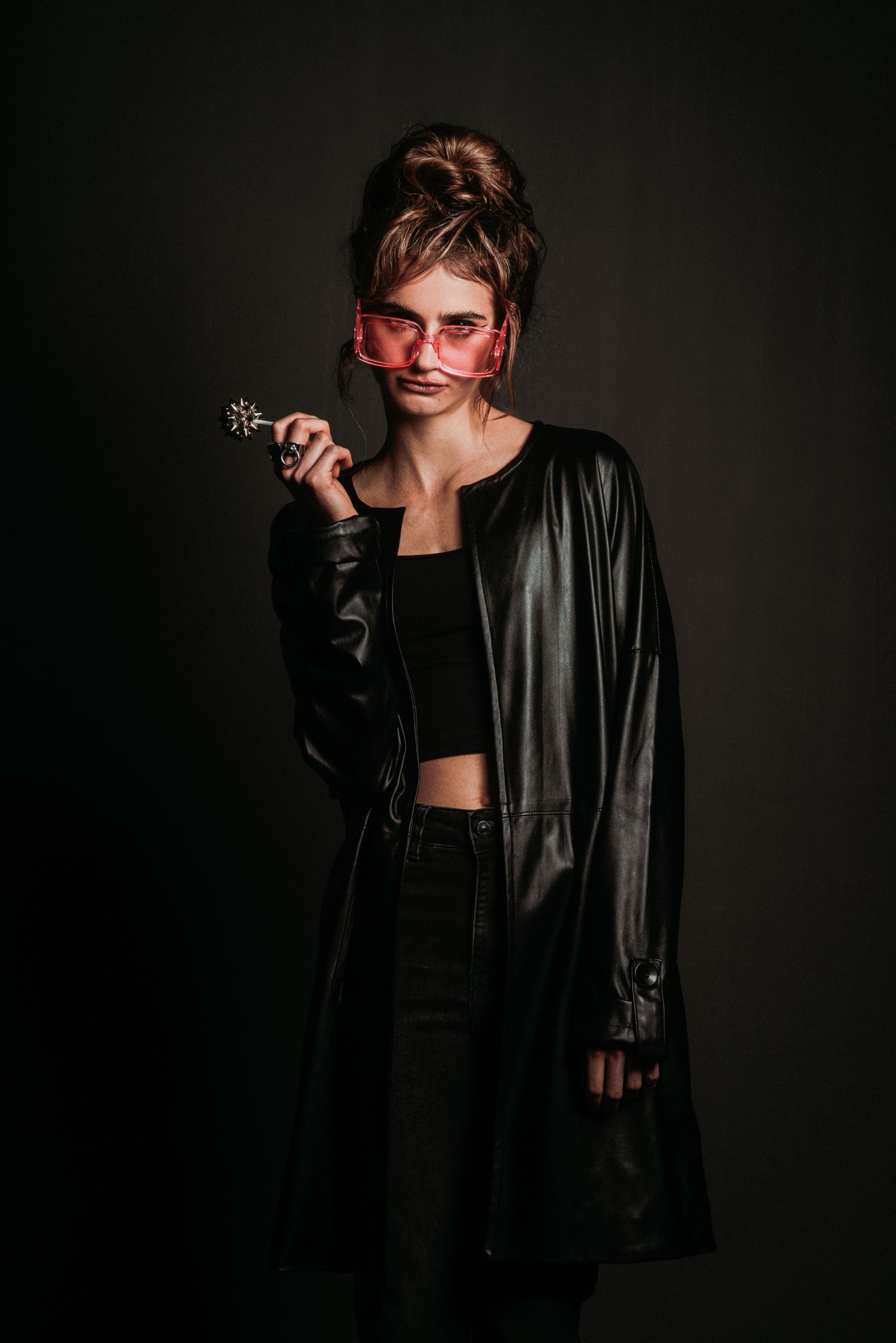 Young woman in black leather coat and pink sunglasses holding a decorative wand against dark background.