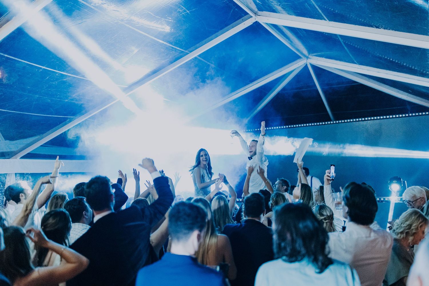 Crowd of wedding guests dance under blue lights and fog in elegant tent reception venue.