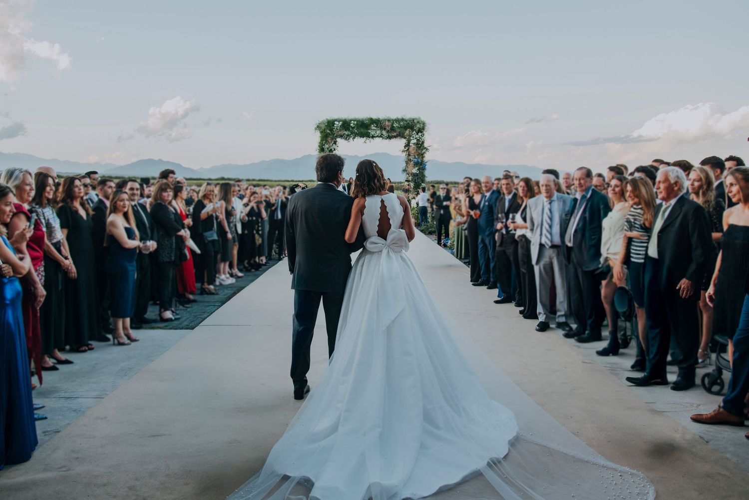 Bride and groom walk down the aisle toward a floral arch, surrounded by wedding guests at an outdoor ceremony.