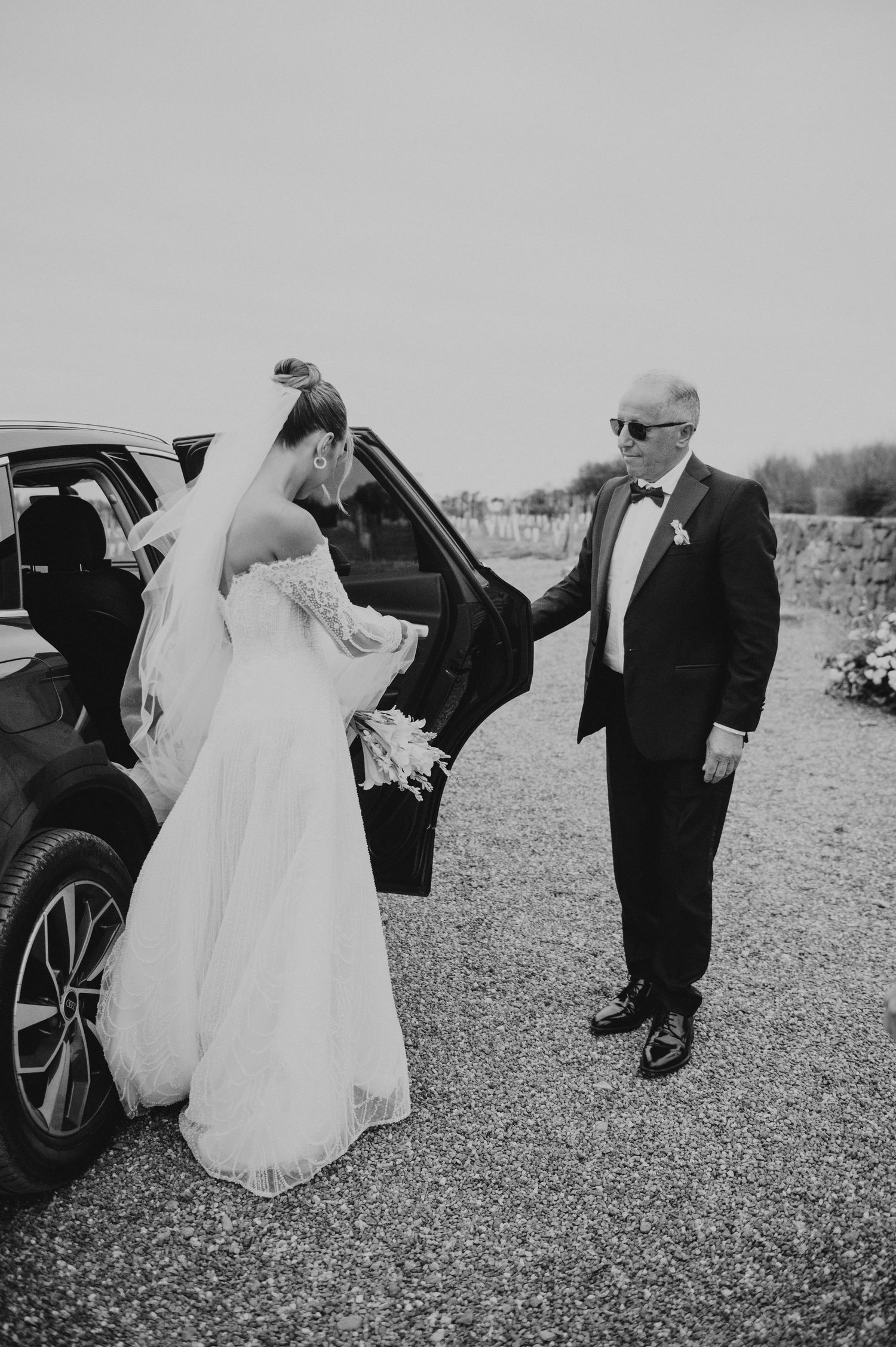 Black and white photo of bride exiting car assisted by older man in tuxedo at wedding venue.