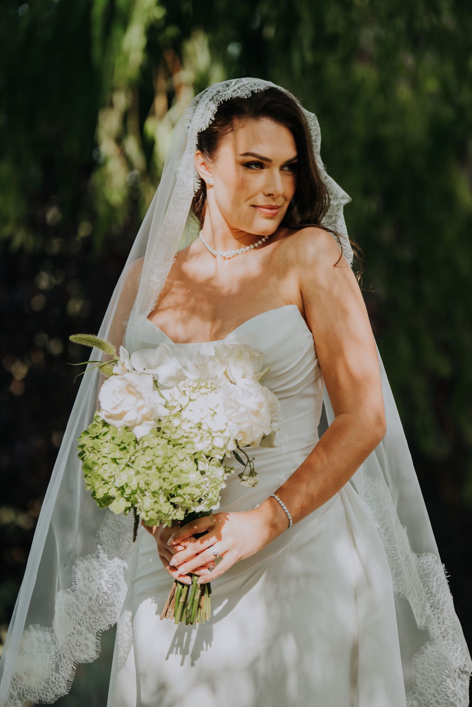 Bride in strapless white gown and lace veil holds white rose bouquet outdoors.