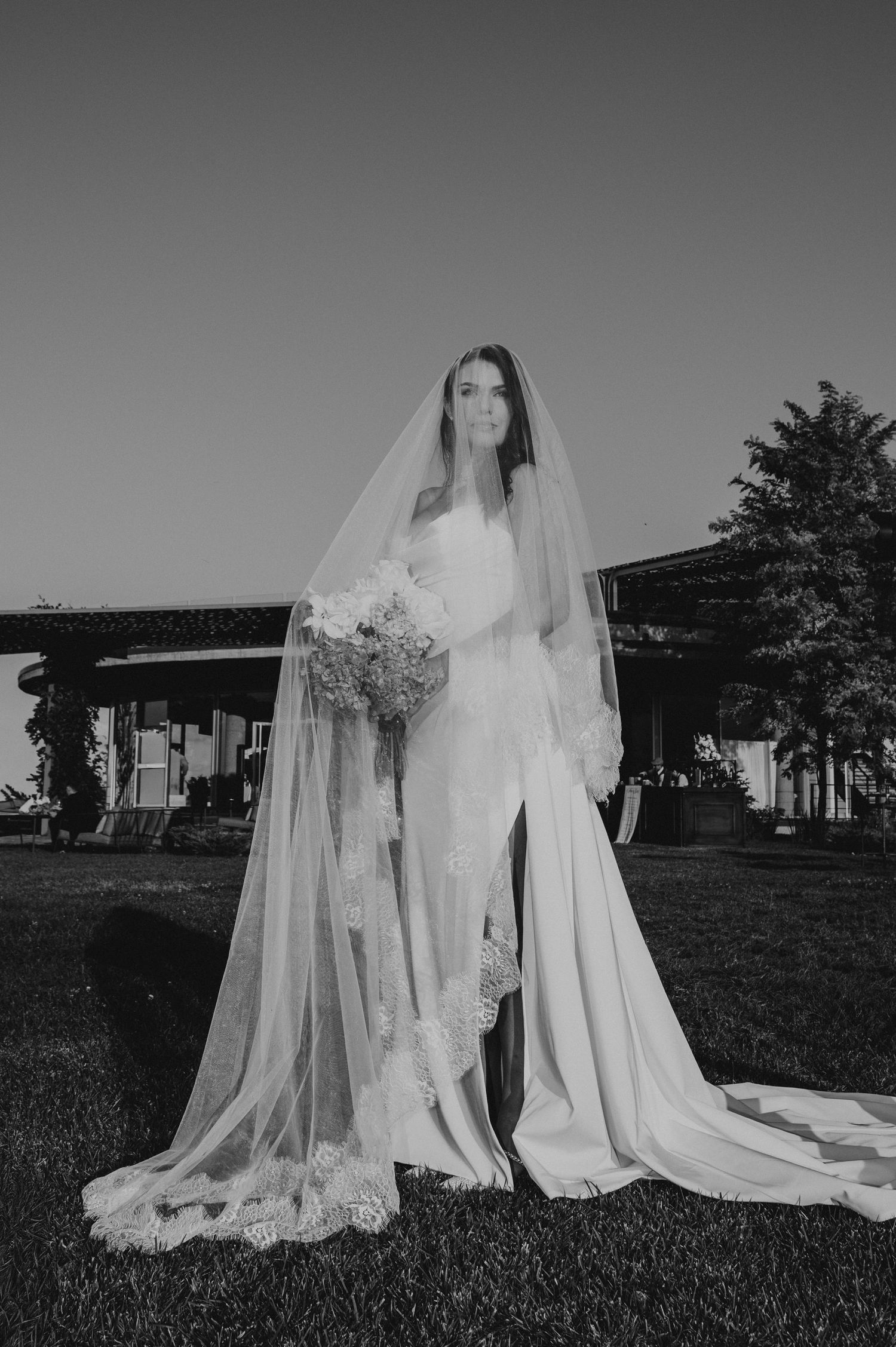 Black and white full-length portrait of bride in lace veil holding bouquet outdoors at dusk.