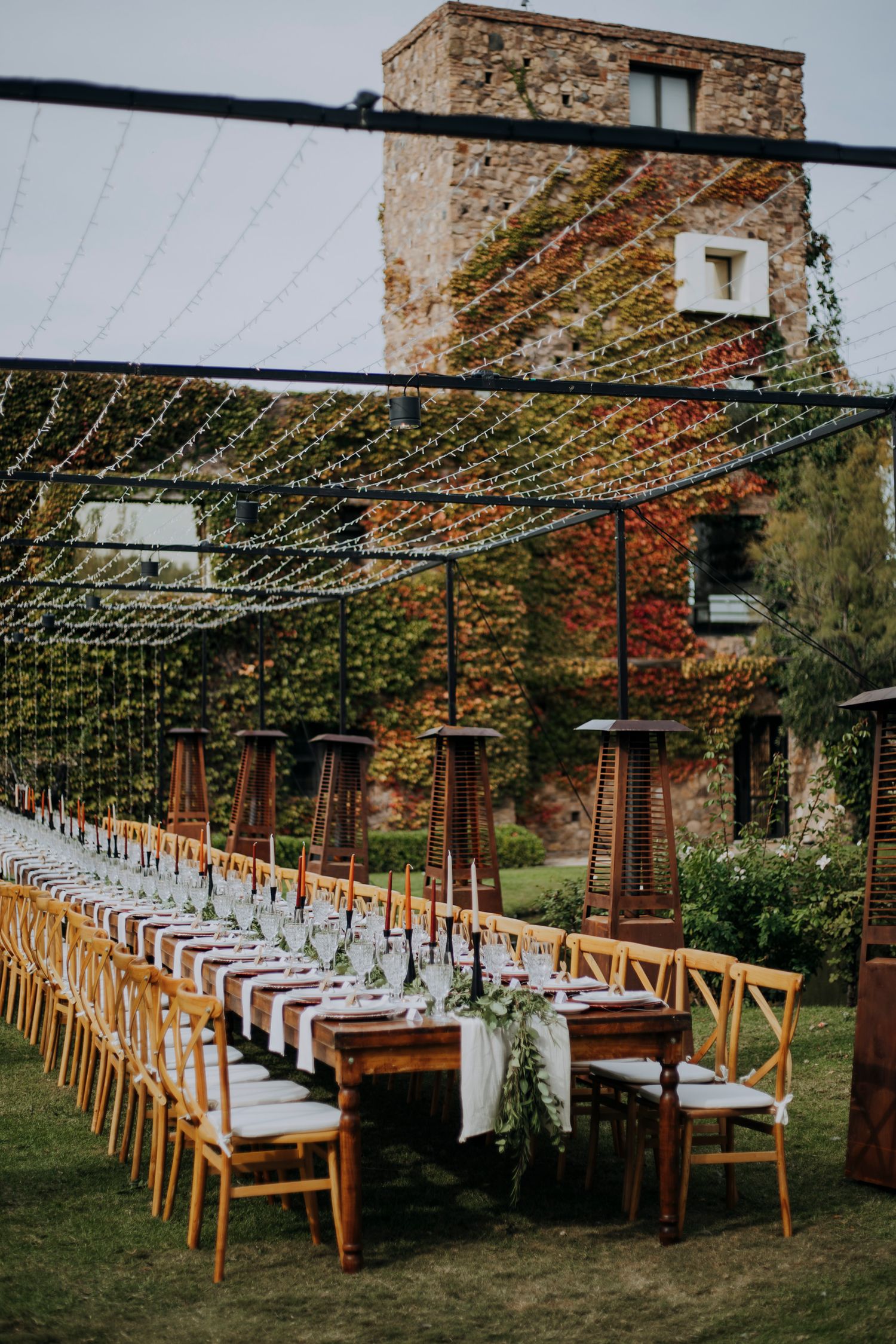 Elegant outdoor wedding reception long table with greenery runner beneath string lights and ivy-covered stone tower.