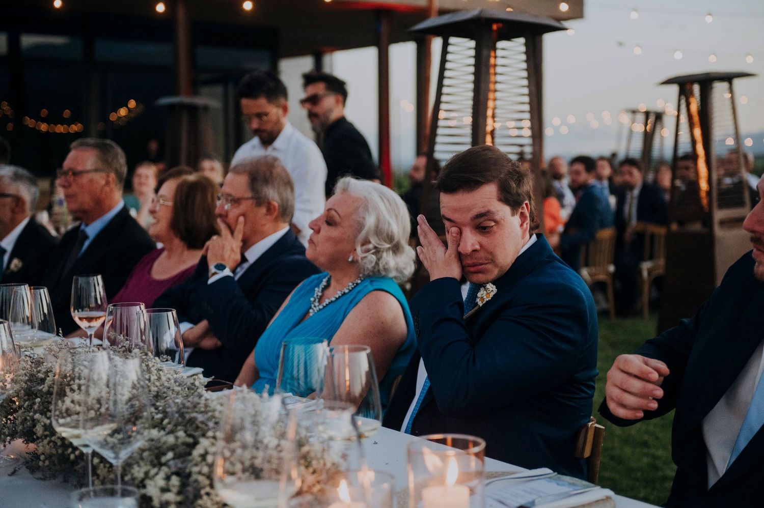 Wedding guests seated at reception dinner table with candles and floral centerpieces at dusk.