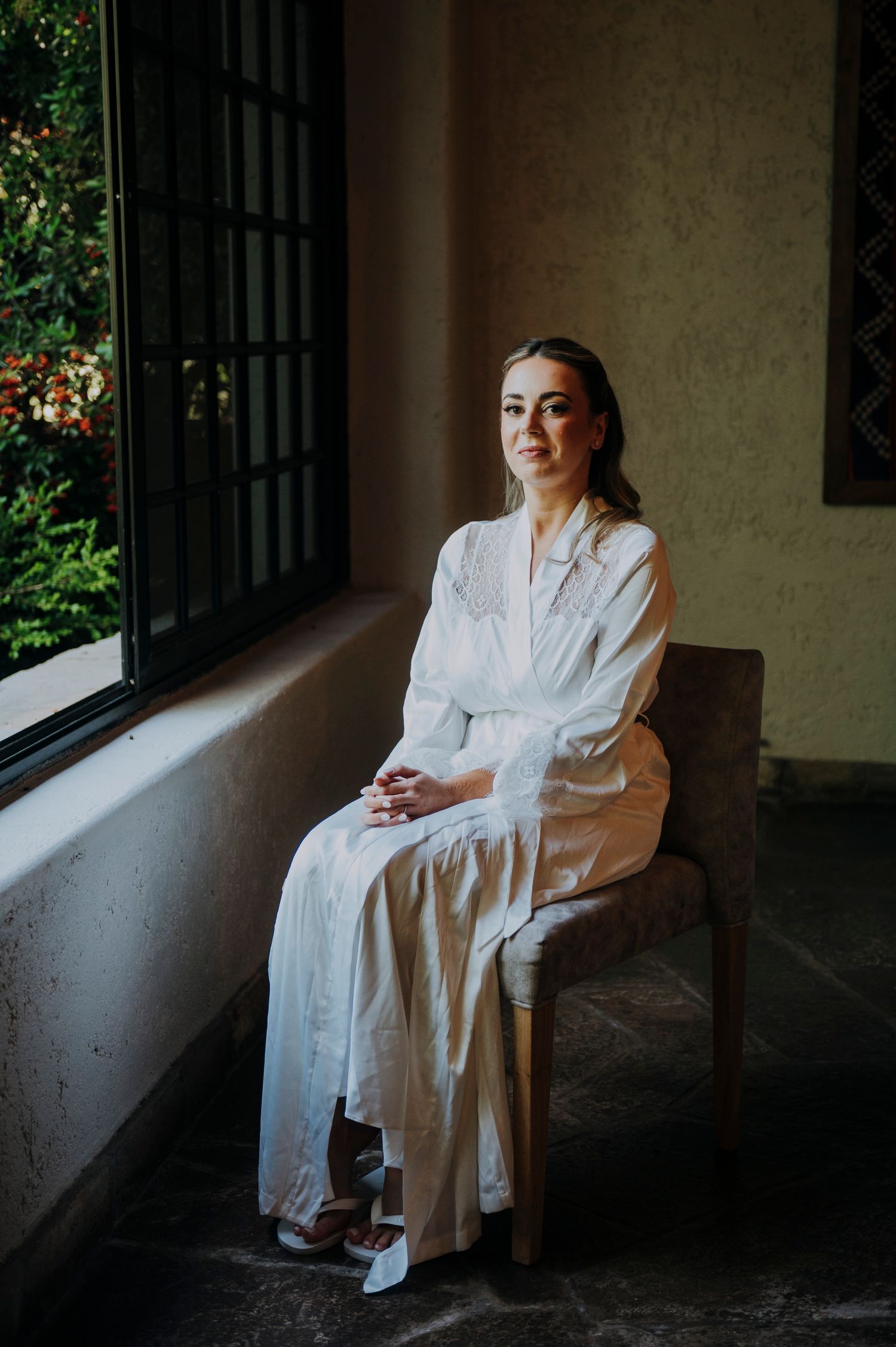 Bride in white satin robe sitting peacefully by sunlit window before wedding ceremony.