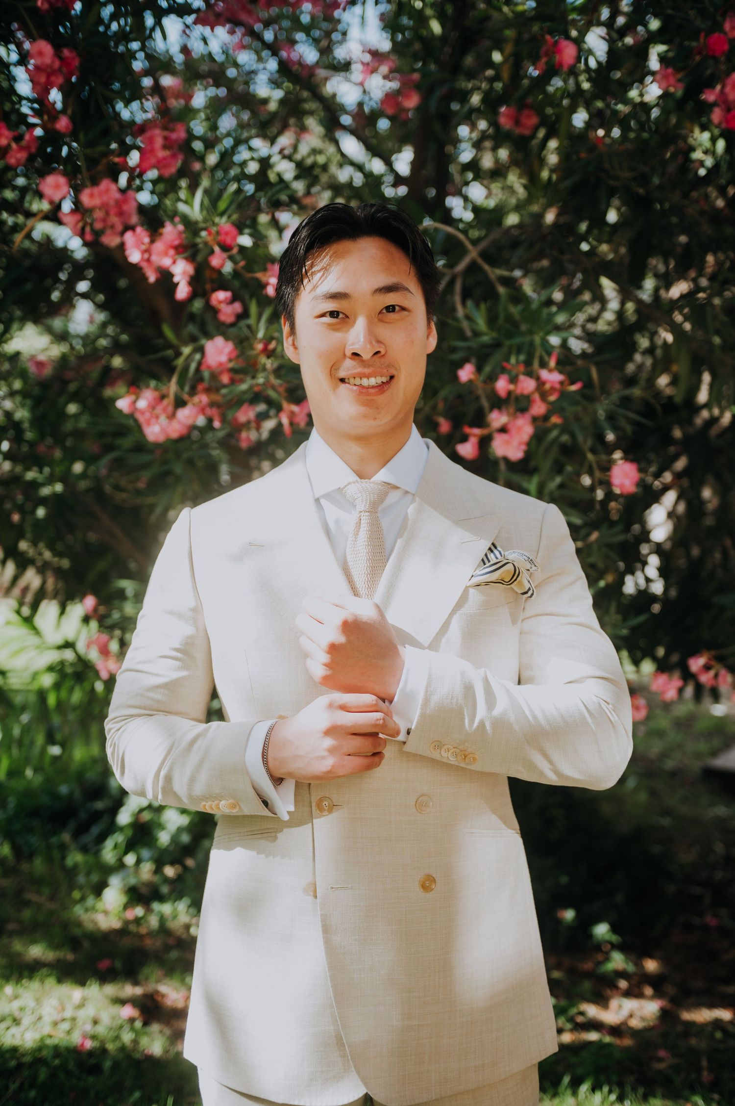 Groom in cream double-breasted suit adjusting cufflinks outdoors with pink flowering shrubs in background.
