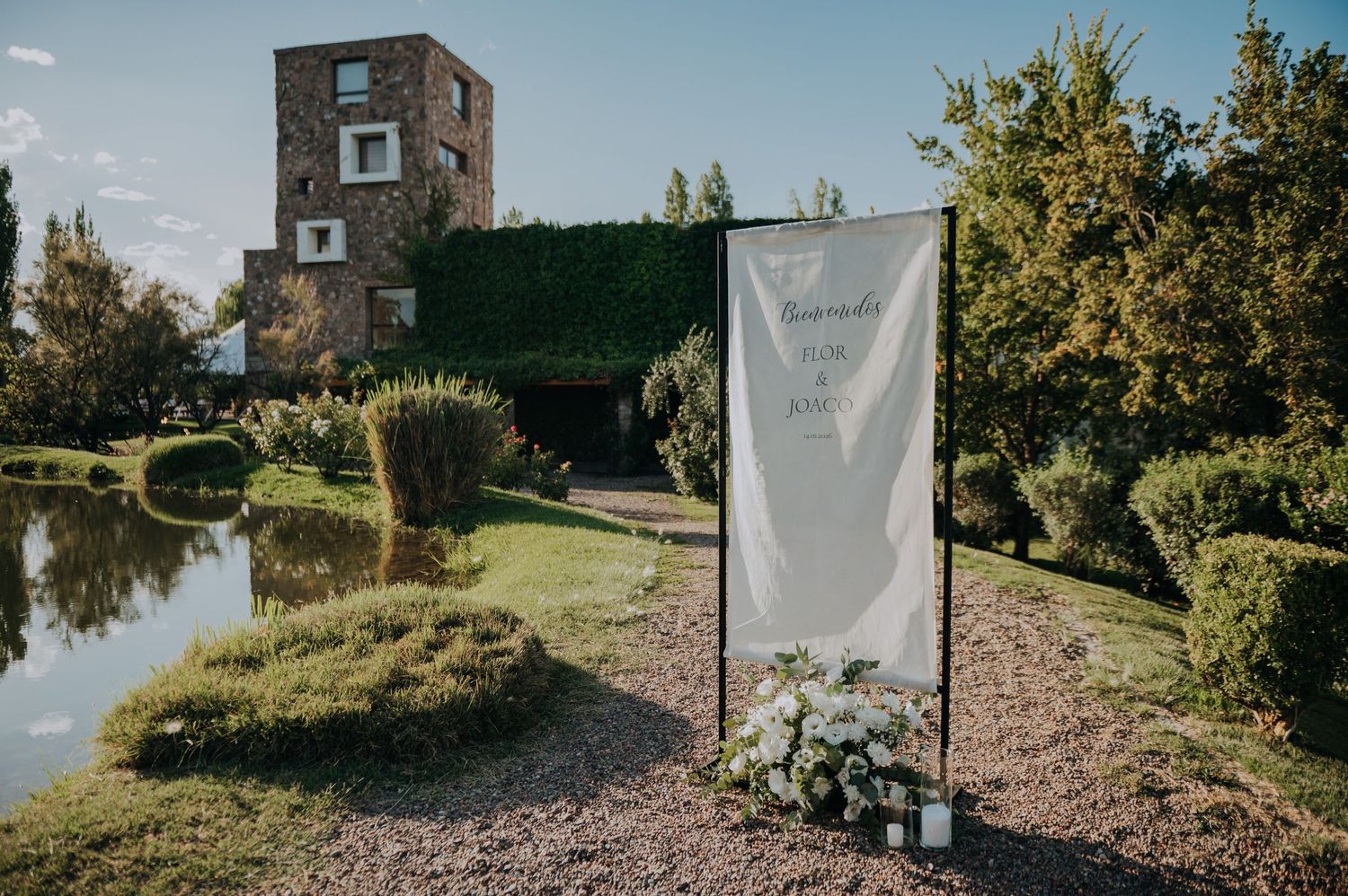Elegant outdoor wedding welcome sign beside a pond with manicured hedges and stone building backdrop.