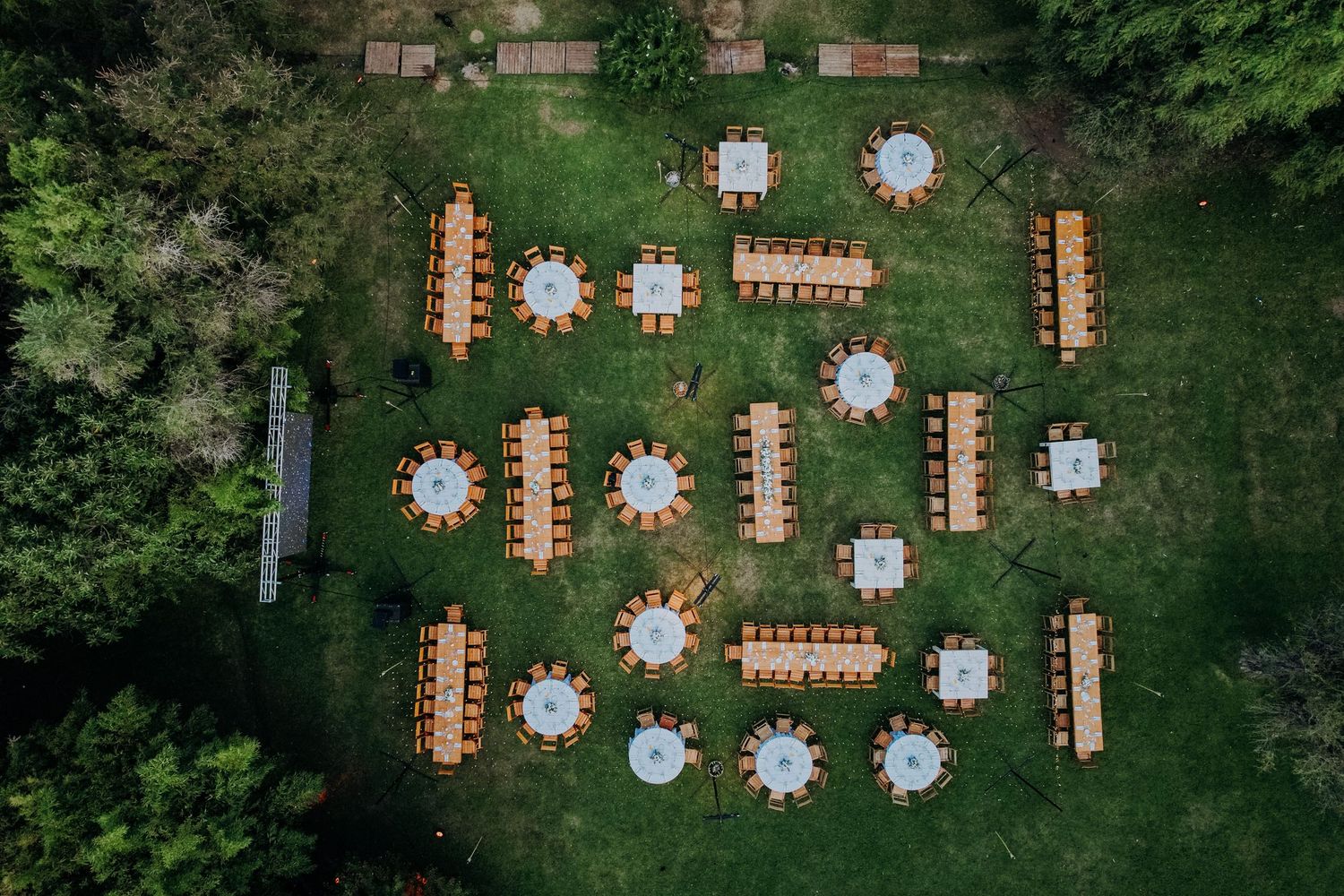 Aerial view of outdoor wedding reception with round and rectangular tables arranged on green lawn surrounded by trees.