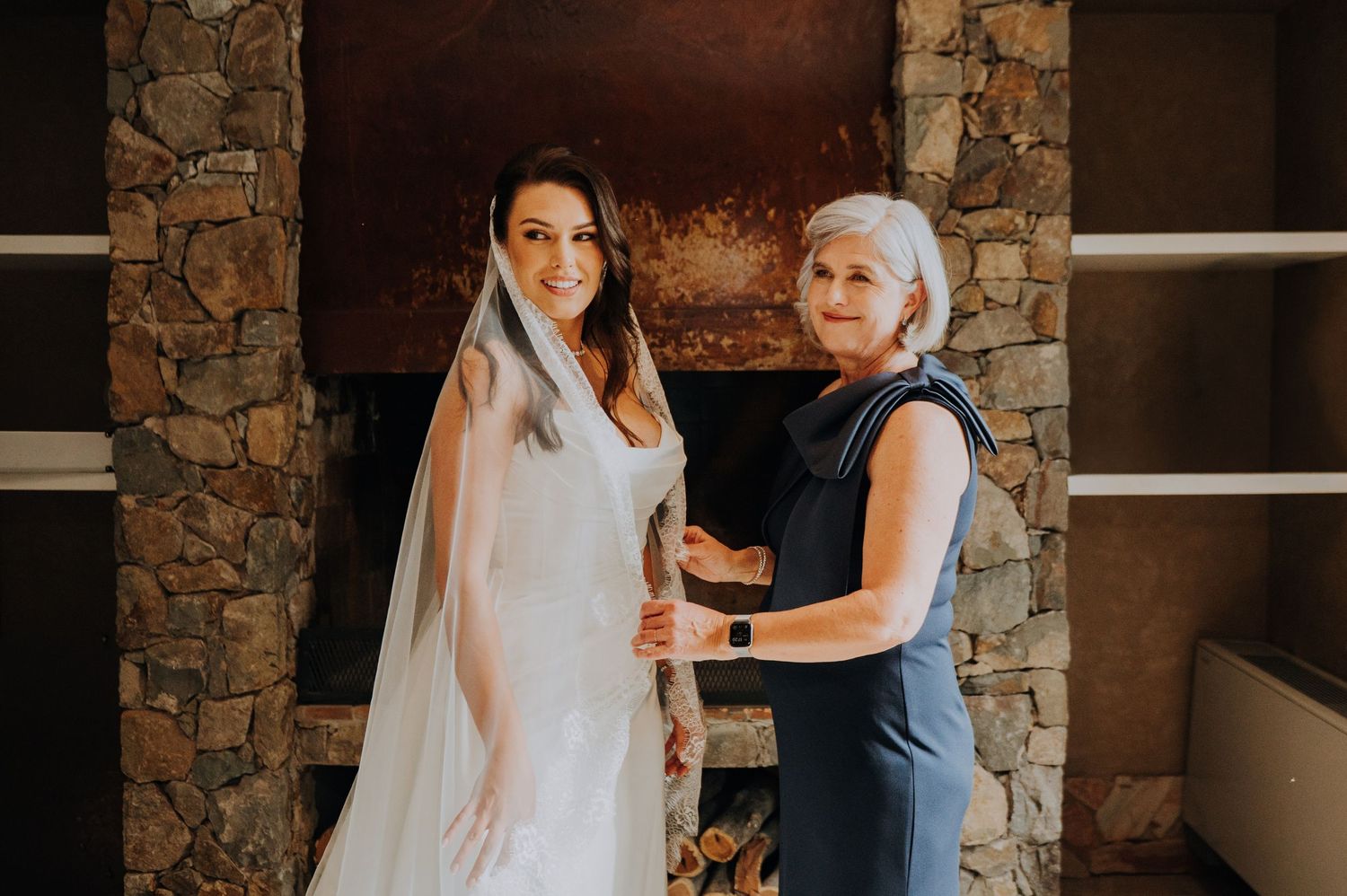 A smiling bride in a white gown and veil stands with an older woman near a stone fireplace.