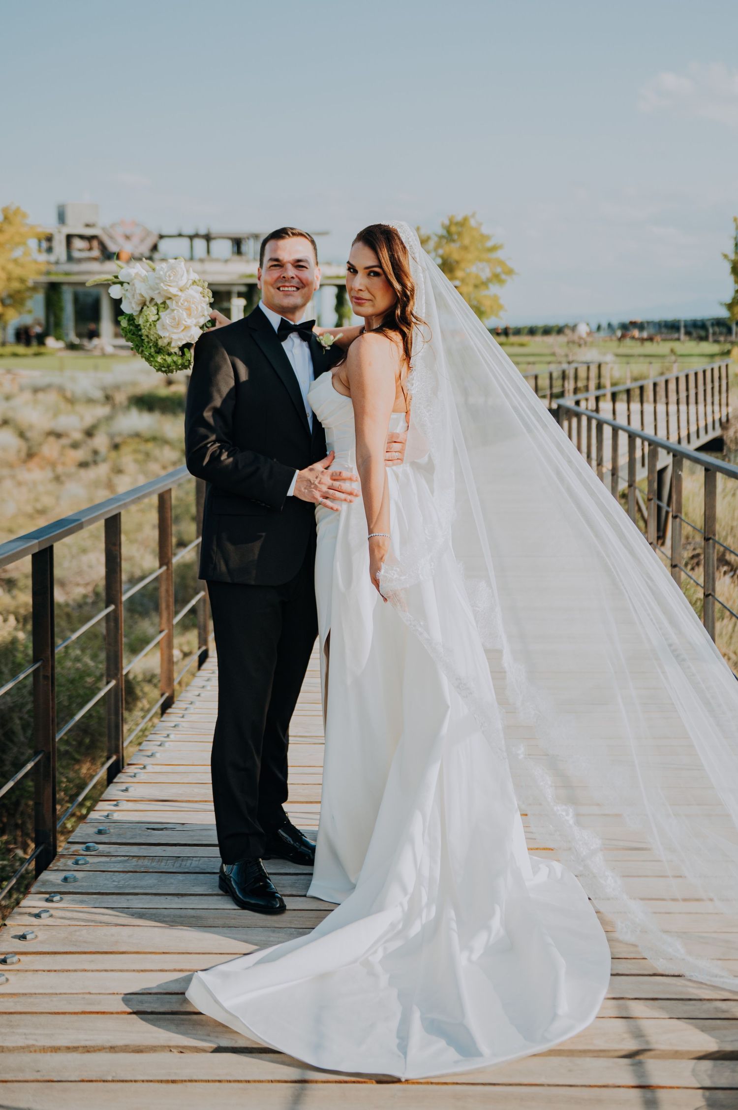 Bride in flowing white gown with long veil and groom in black tuxedo posing on wooden bridge outdoors.