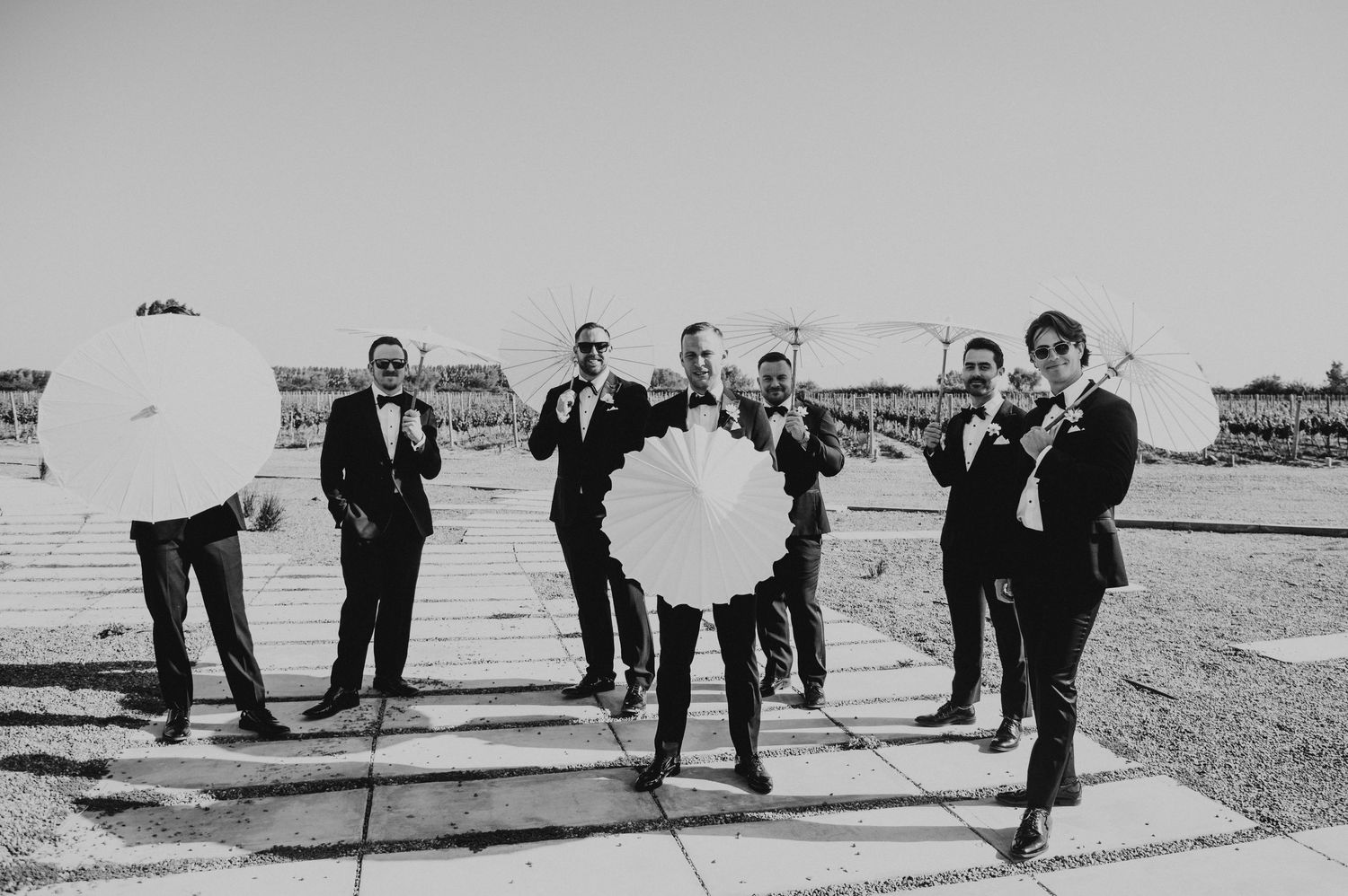 Groomsmen in black tuxedos hold white parasols and bouquets in a fun black and white outdoor photo.