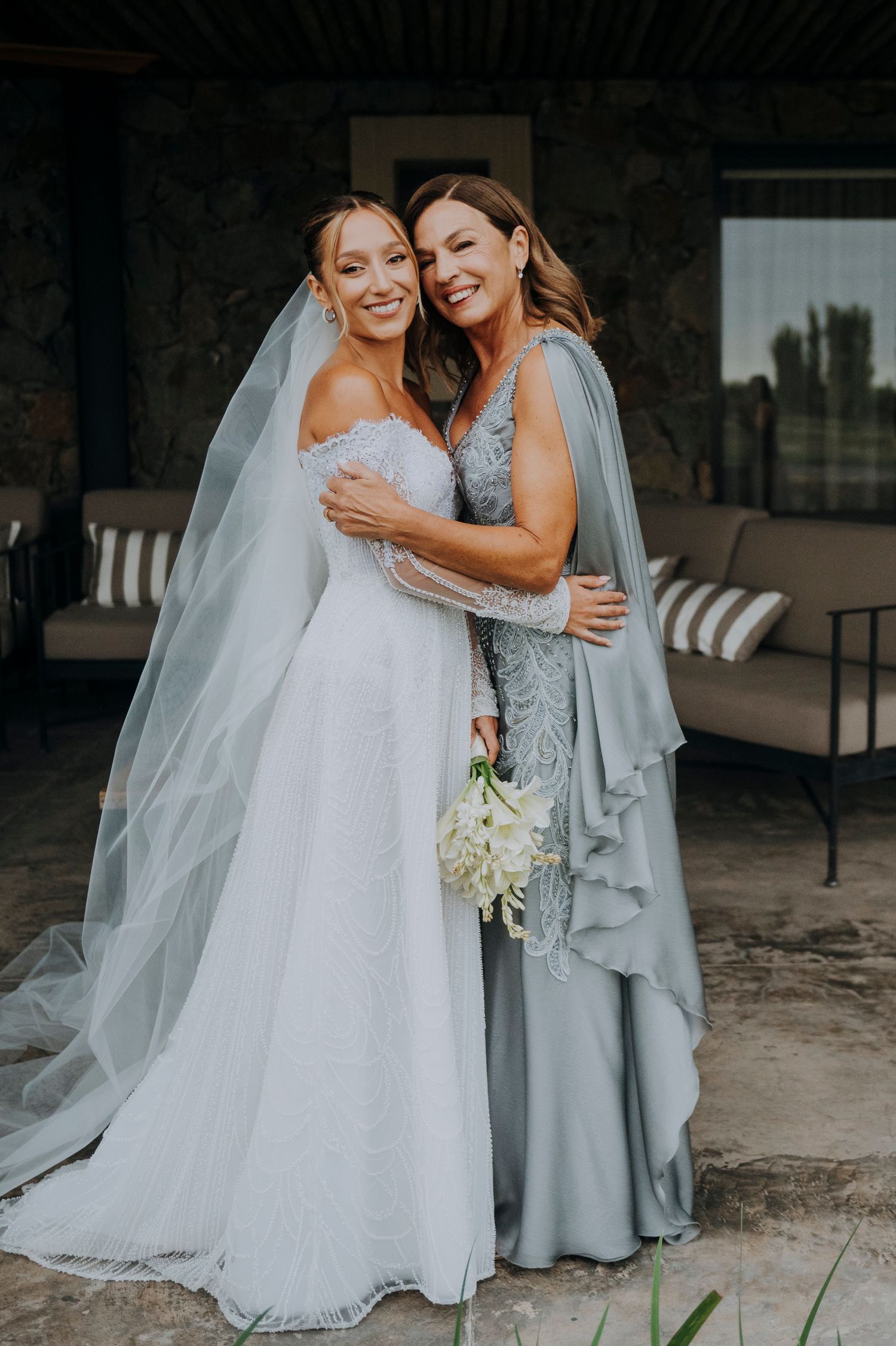 Bride in white off-shoulder gown and veil poses with woman in sage green beaded dress holding white bouquet.