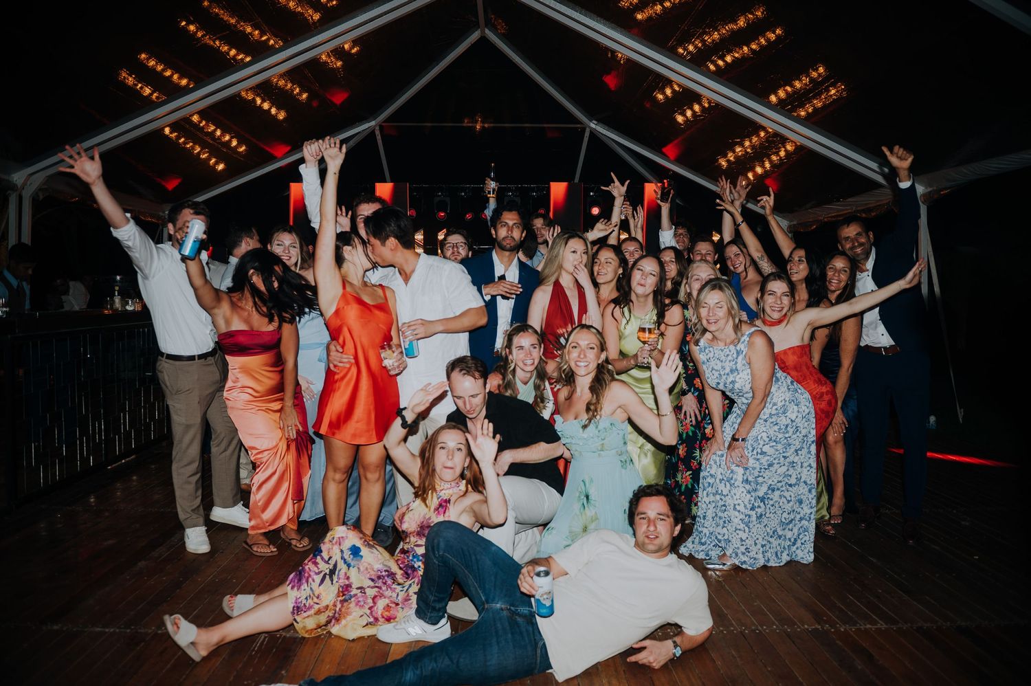 Joyful wedding reception guests posing together on dance floor under warm string lights at lively celebration.