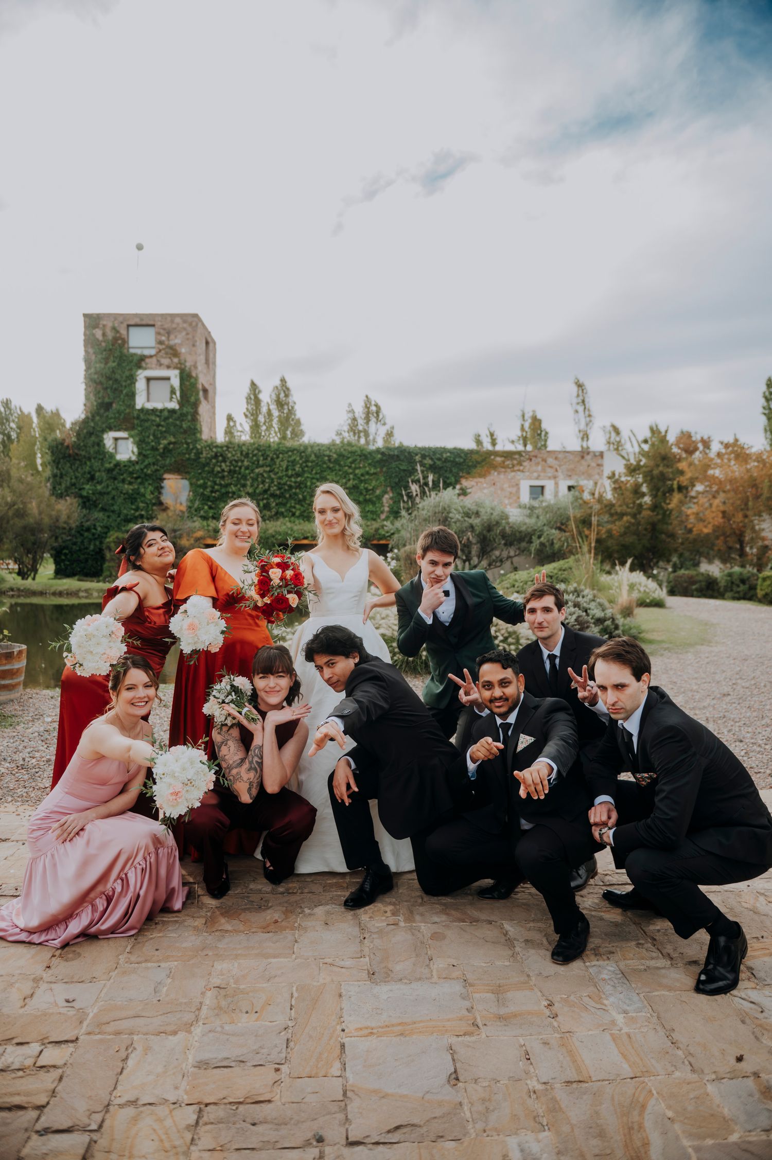 Wedding party posing playfully outdoors with bridesmaids in red and pink dresses and groomsmen in black suits.