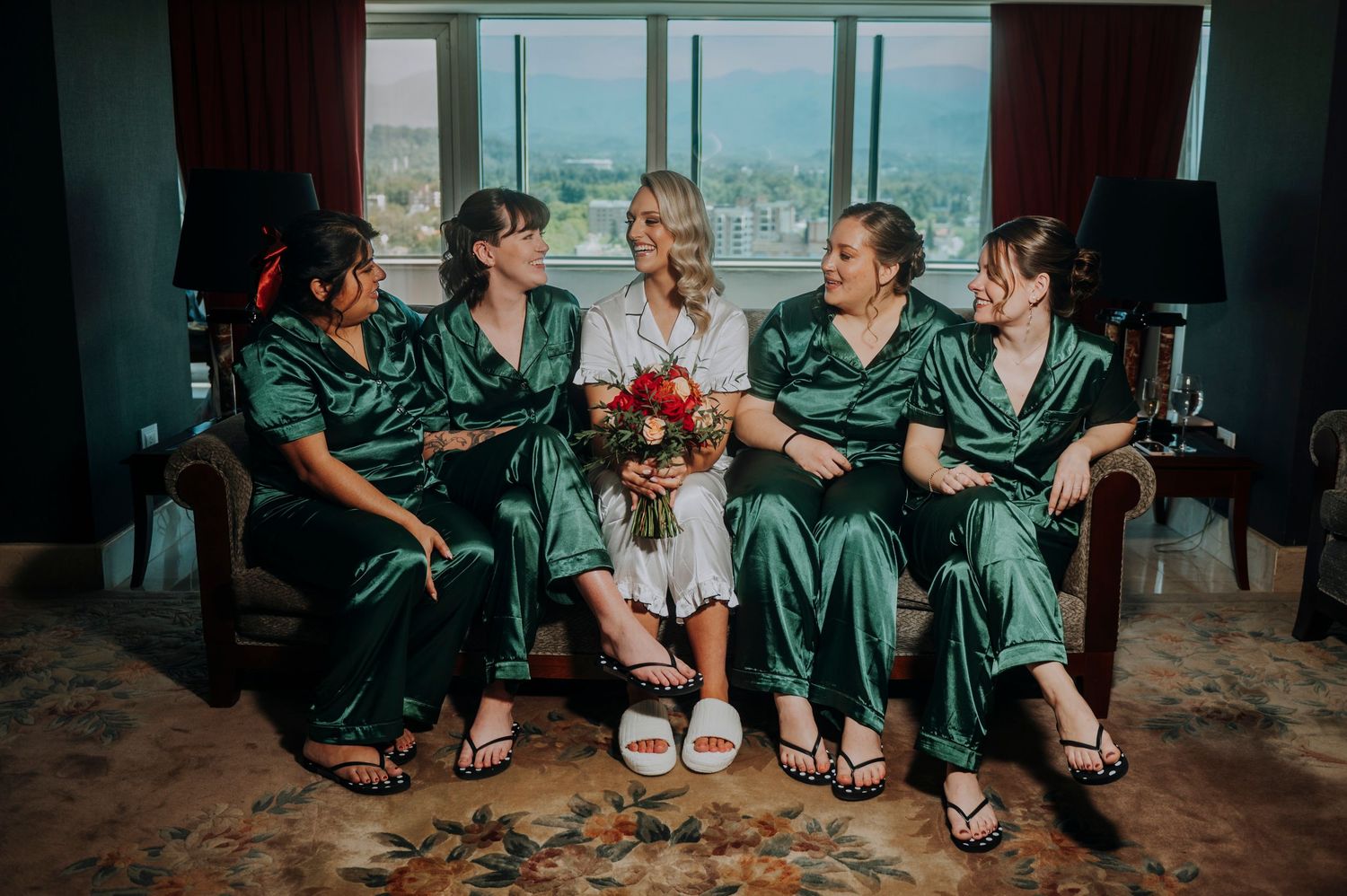 Bride in white robe sits with four bridesmaids in green satin pajamas holding a bouquet in a hotel room.