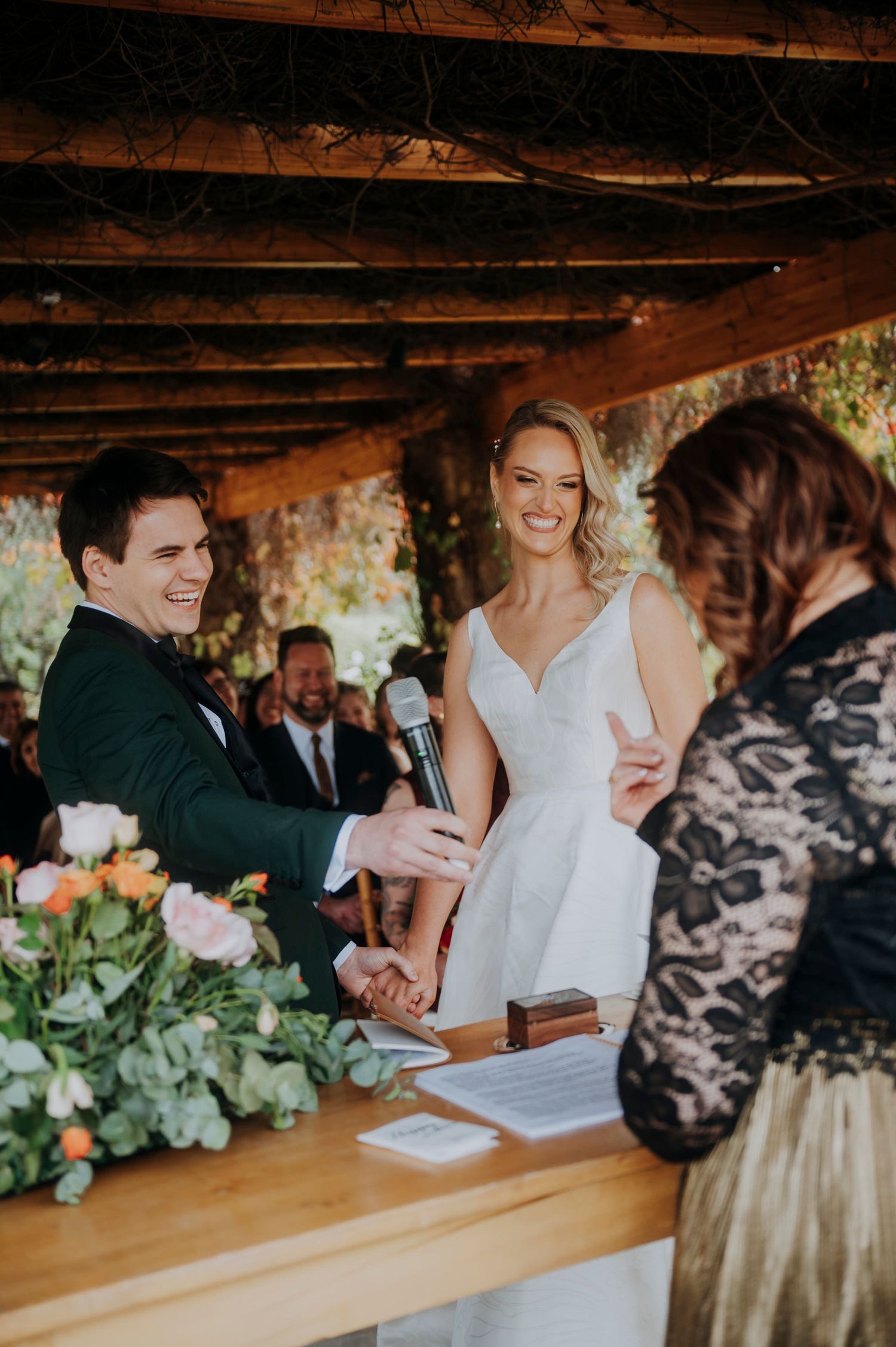 Bride and groom laugh together at altar with officiant during outdoor wedding ceremony.