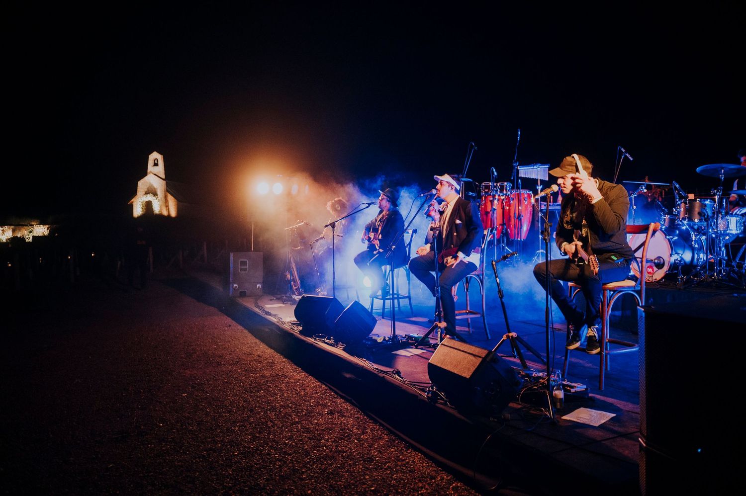 Band performing on outdoor stage at night with blue lights and illuminated church tower in background.