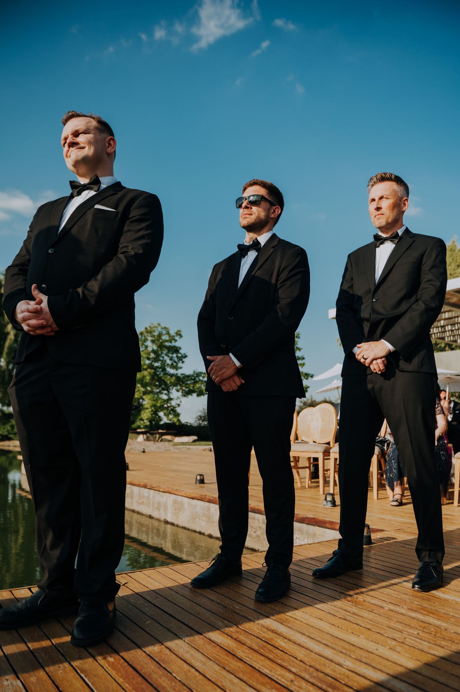 Three groomsmen in black tuxedos standing on wooden deck during sunny outdoor wedding ceremony.