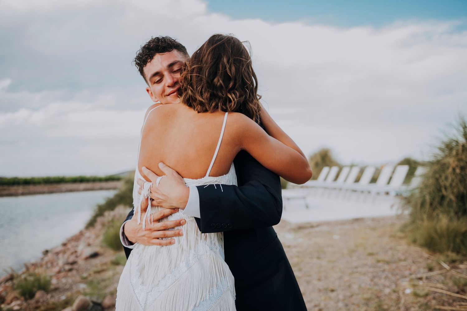 Bride and groom sharing a heartfelt embrace outdoors near a scenic waterway on their wedding day.