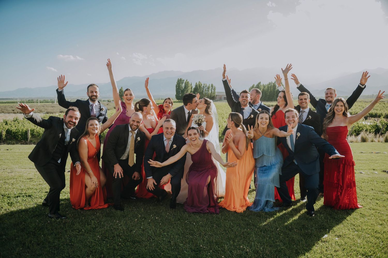 A joyful wedding party group photo with colorful dresses and raised hands in a vineyard setting.