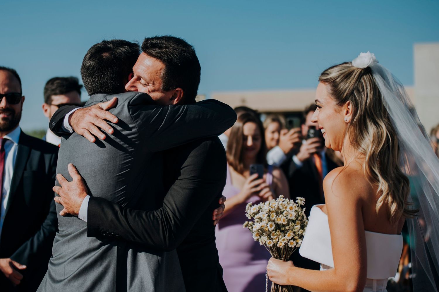 Groom embraces a guest warmly as the bride in white dress and veil smiles during outdoor wedding ceremony.